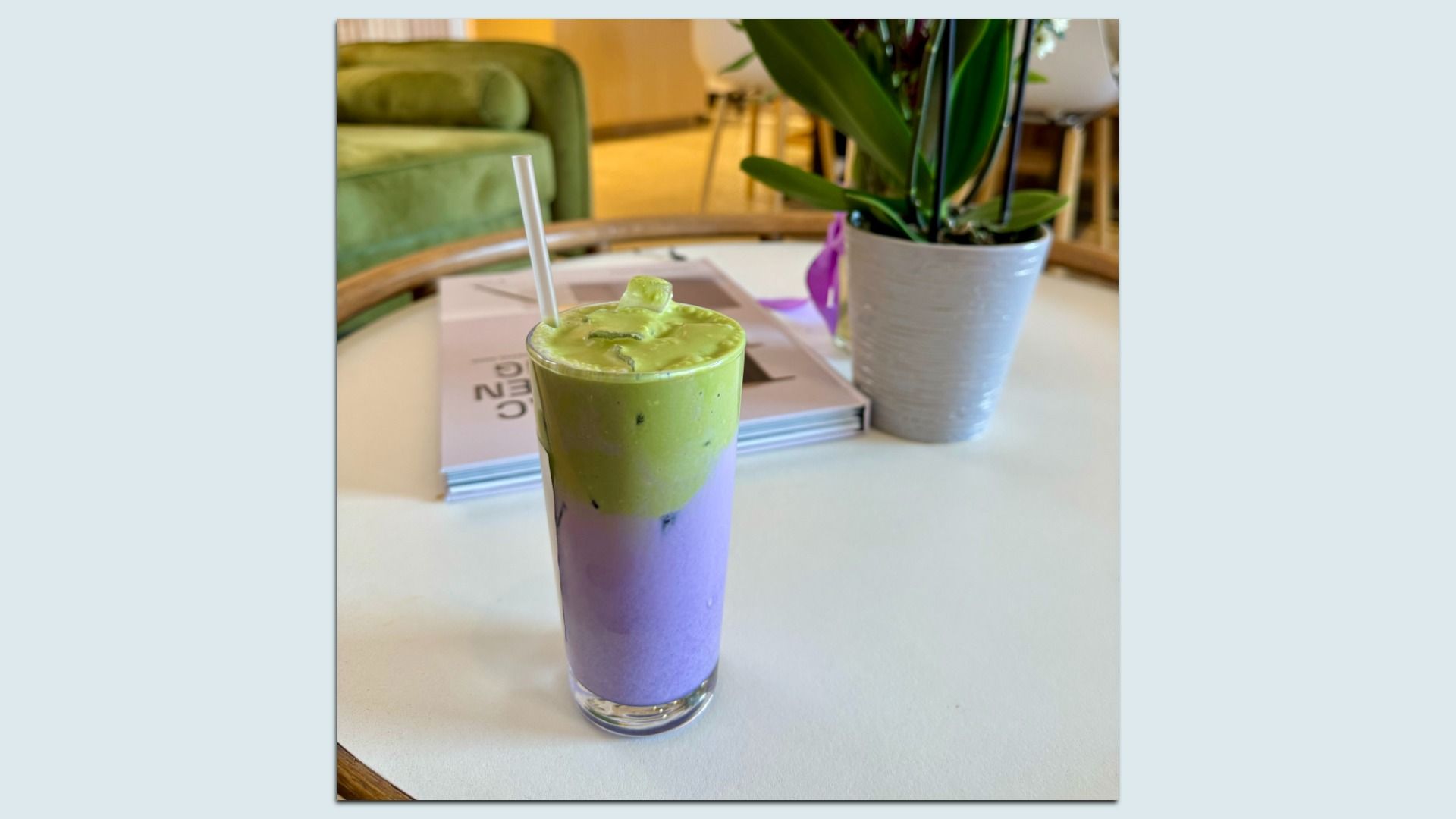 Tall glass on a white table with a layered iced drink, green on top and purple at the bottom, with a white straw. A potted plant and a magazine are in the background.