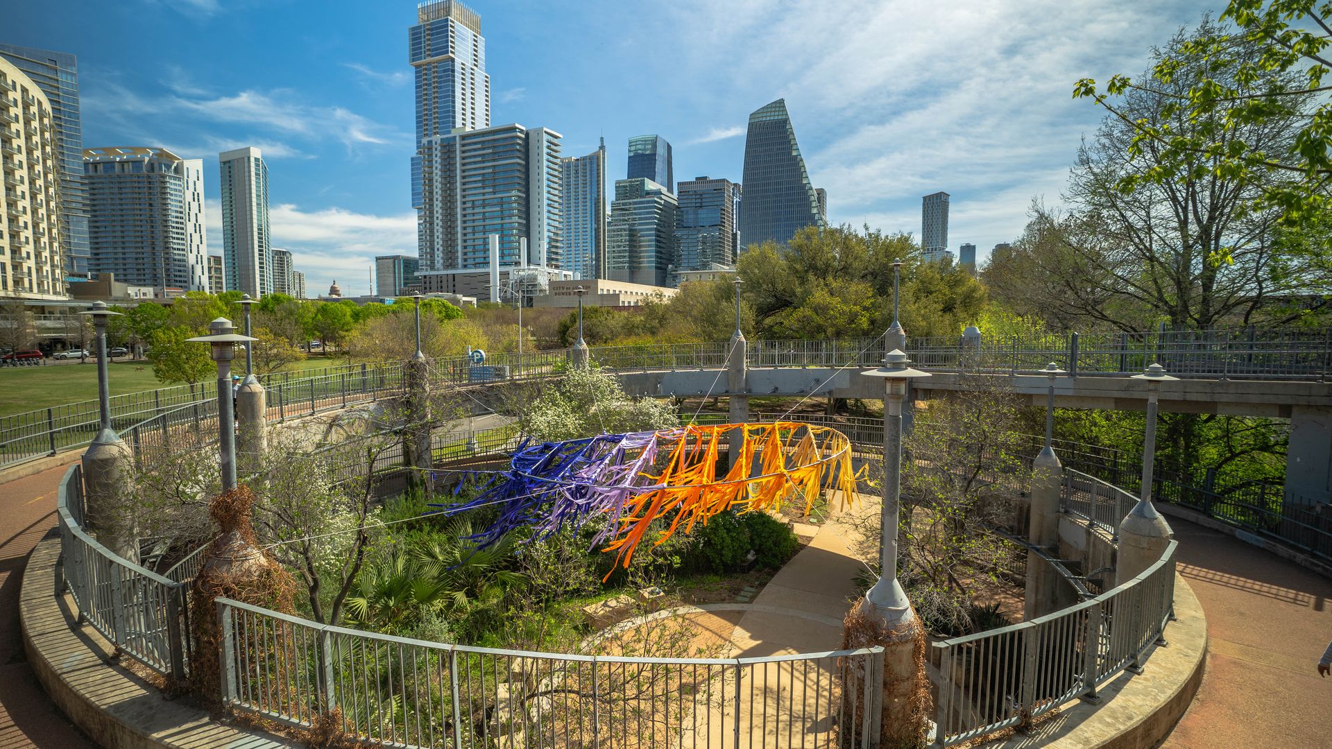 Circular garden with a colorful ribbon sculpture (blue, purple, orange) in a city park; metal railing surrounds it. Towering skyscrapers rise in the background against a blue sky.