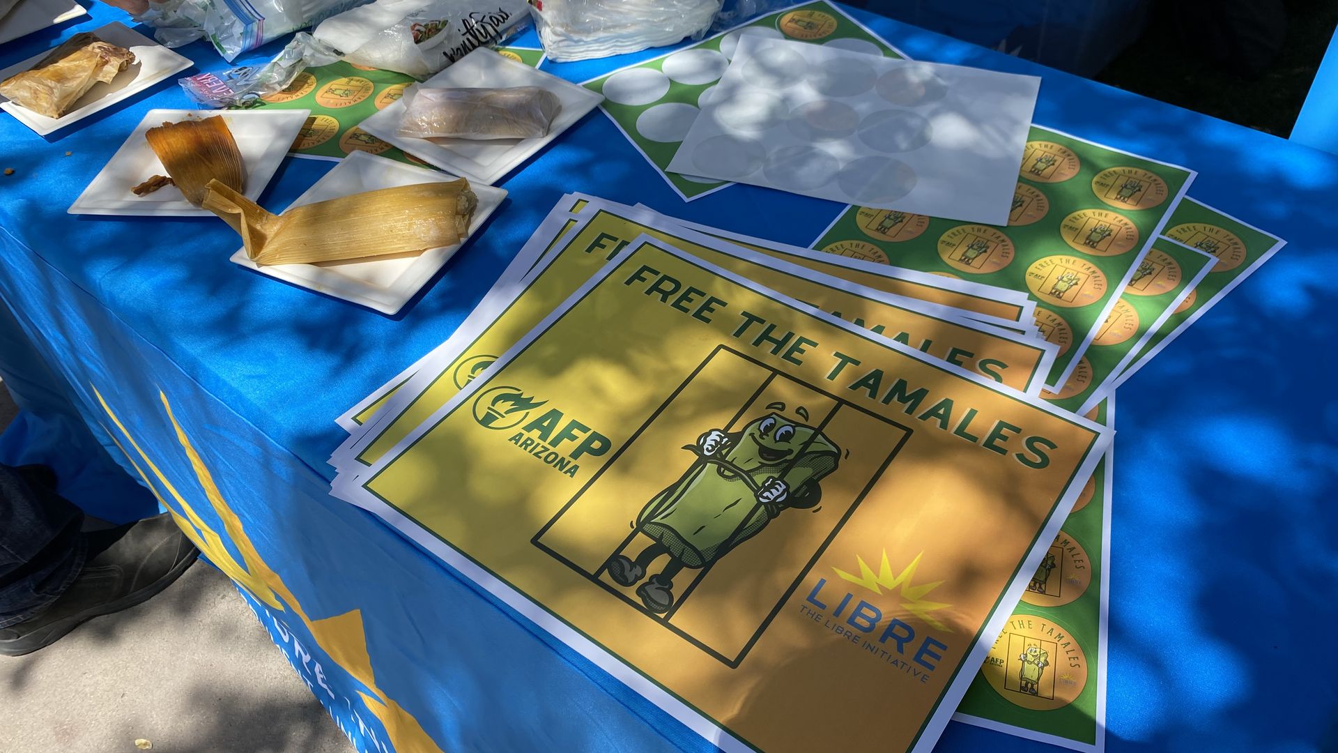 A table filled with tamales, sheets of stickers and yellow signs that read Free The Tamales.