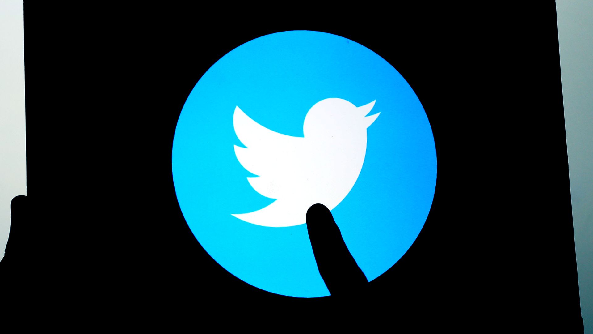 A photo illustration of a hand in silhouette pointing at the Twitter logo prominently displayed on a tablet screen.