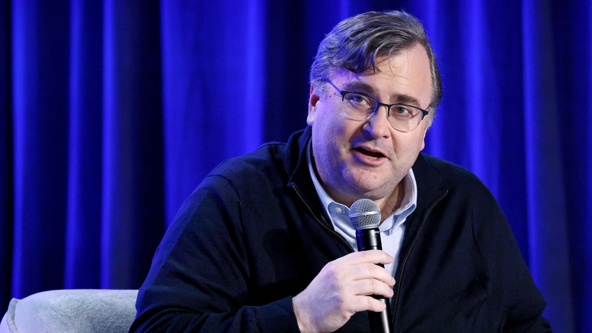 Reid Hoffman holding a microphone in a sweater over a button down sitting in a chair in front of a curtain