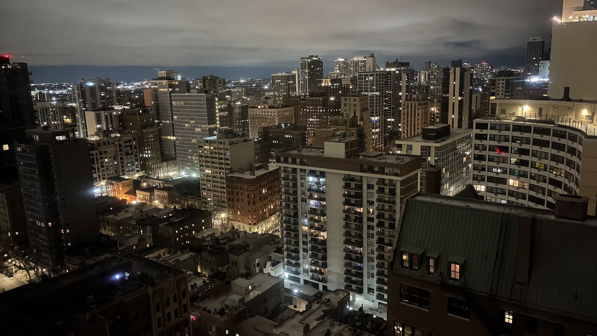 Nighttime cityscape from a rooftop: dense cluster of illuminated high-rise buildings, glowing streetlights, and a cloudy sky, windows lit in warm amber and cool white.