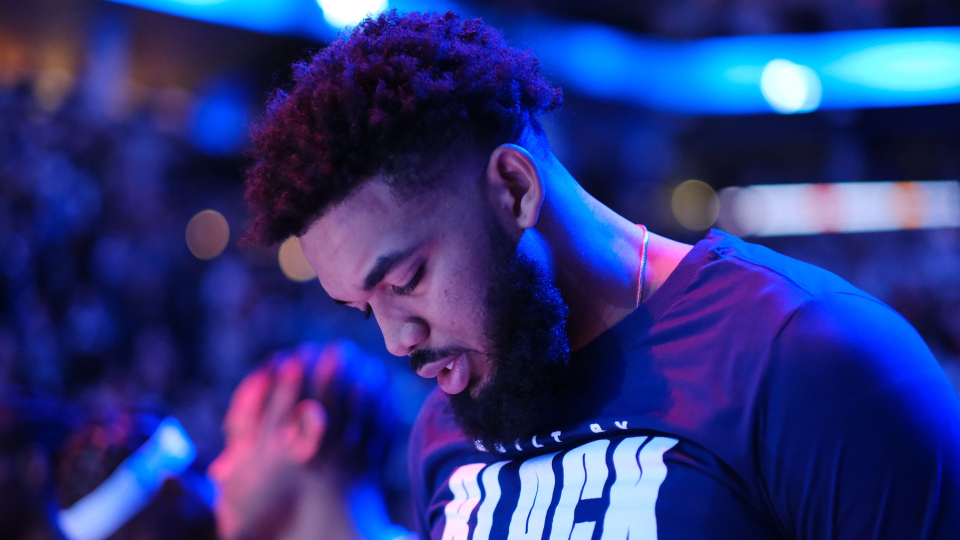 A Black man under blue-tinged lights looks down in a darkened basketball arena.
