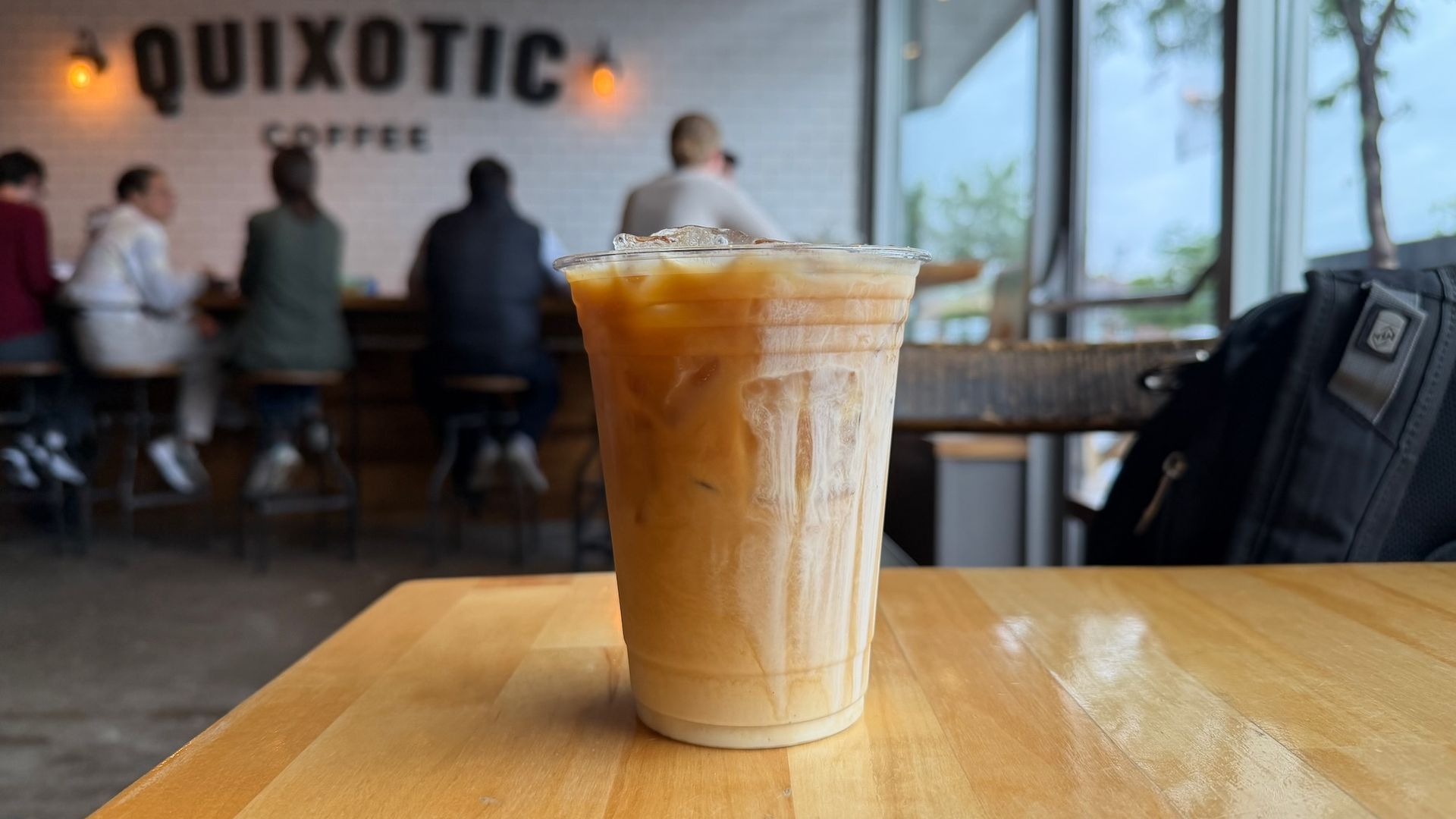 An iced coffee in a clear cup sits on a wooden table in a cafe; blurred customers sit at a counter behind a white tiled wall that reads QUIXOTIC COFFEE, with large windows and trees outside.