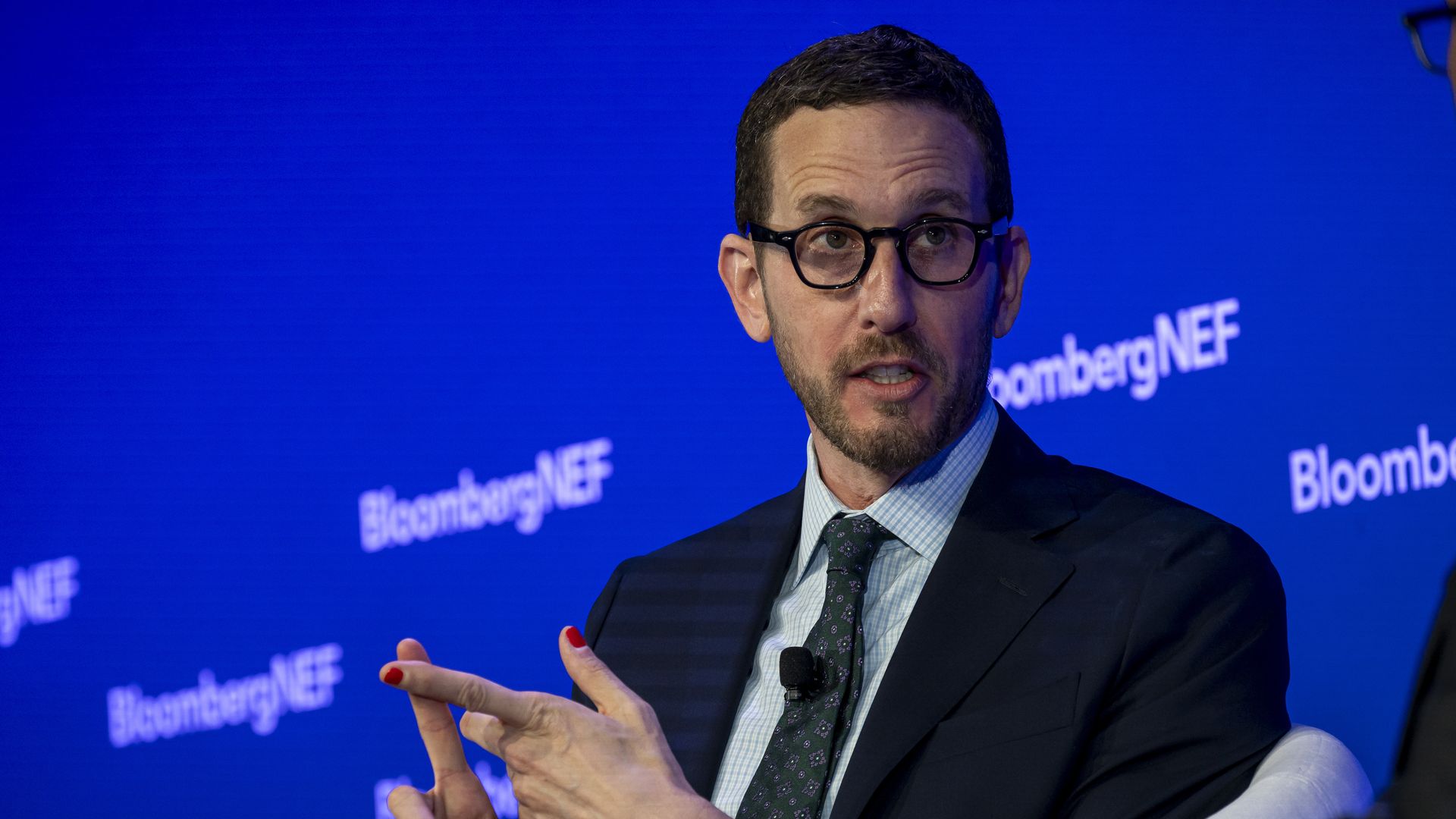 Man in glasses and suit speaking on a panel with a blue BloombergNEF backdrop, gesturing with hands marked by bright red nail polish.
