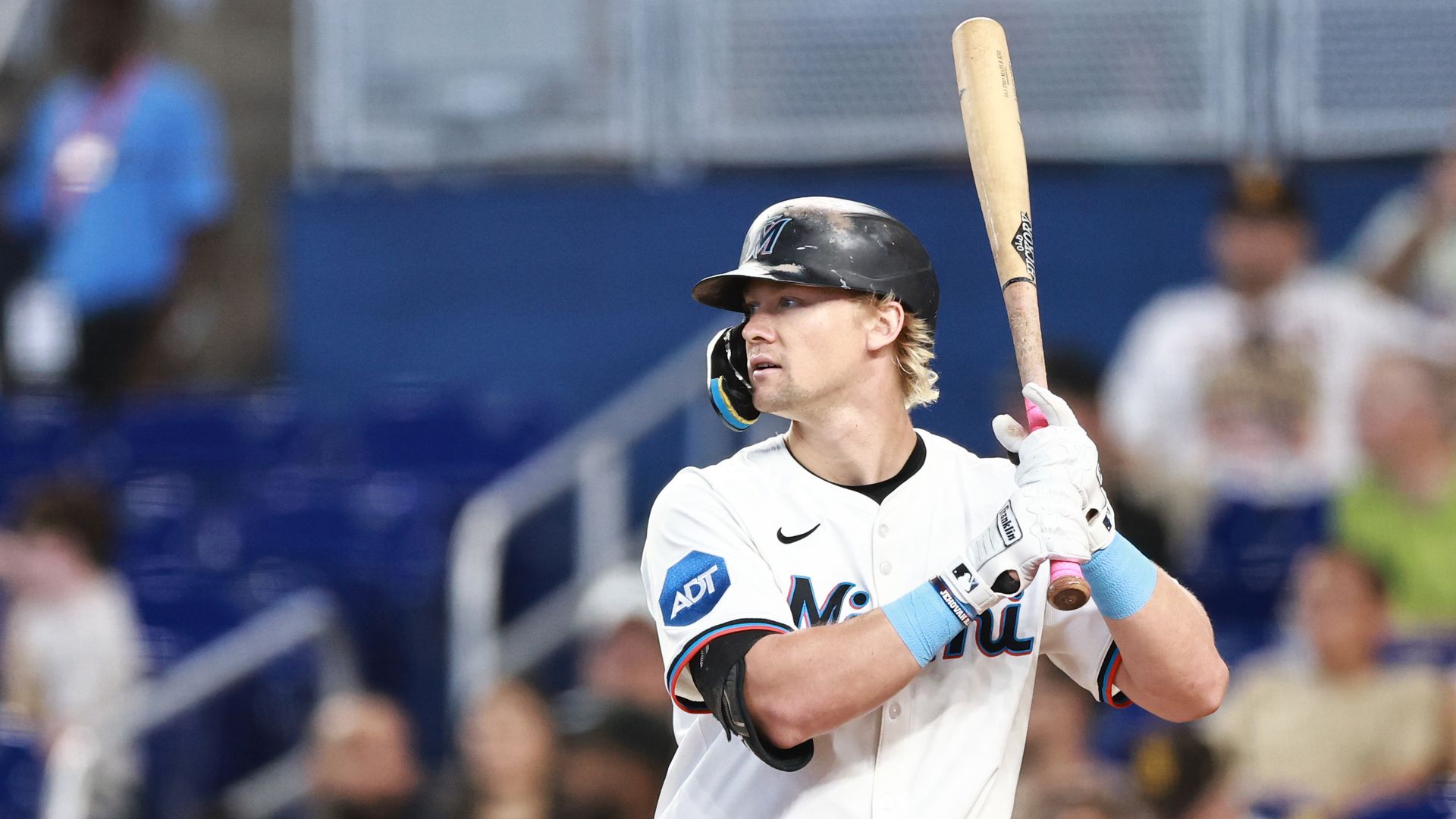 Baseball player in white Miami Marlins uniform with black helmet, holding wooden bat, preparing to bat during a game with blurred spectators in the background.