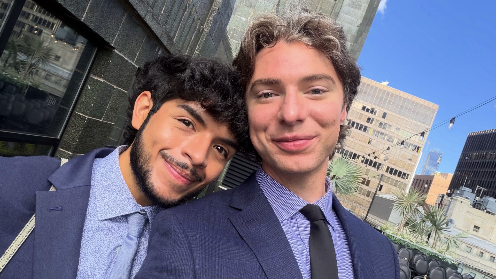 Two young men in navy suits pose for a selfie on a city terrace, smiling. One with dark curly hair, the other with light brown hair. Bright blue sky, modern buildings and string lights behind them.
