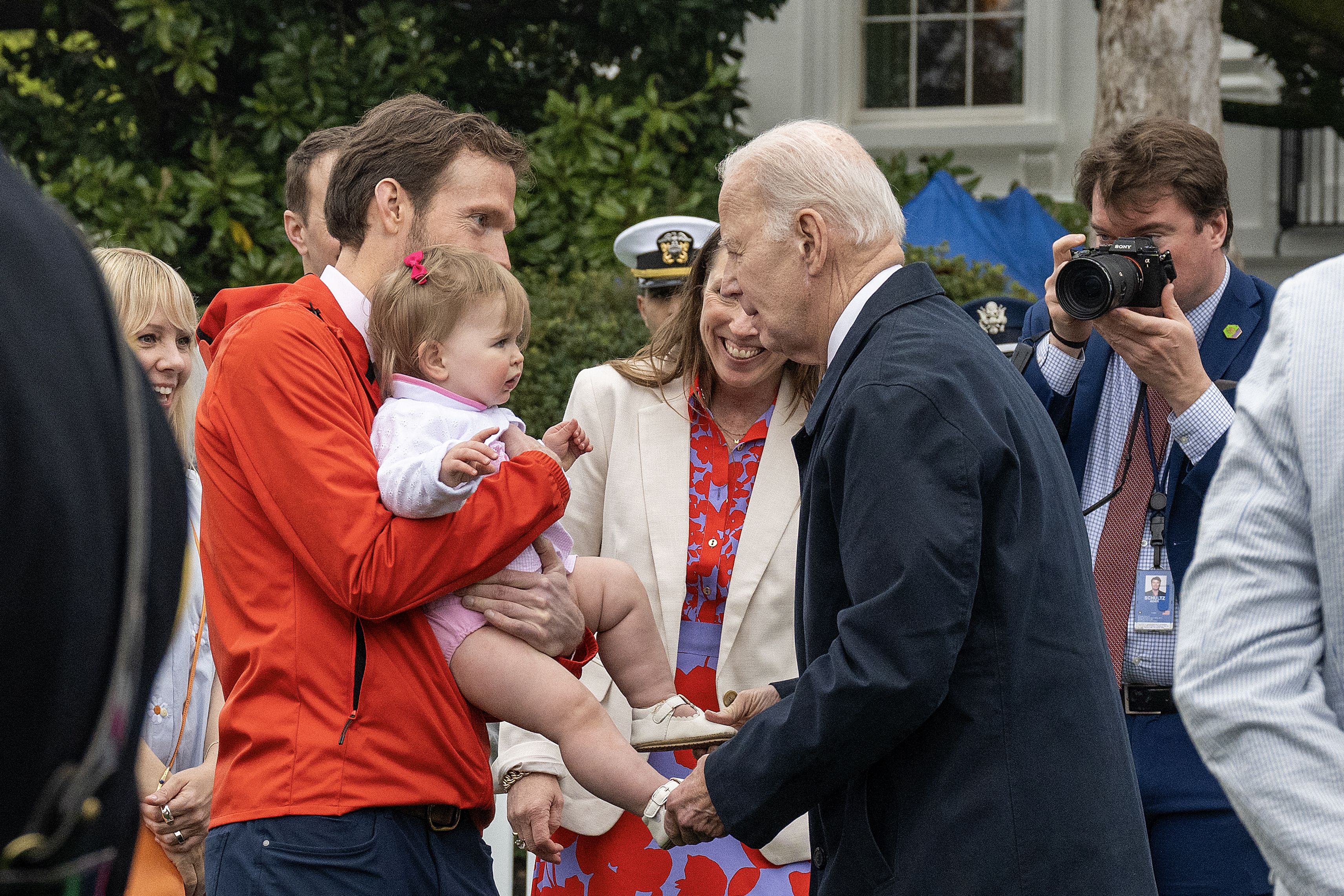Joe Biden greets a baby, being held by an adult with several people surrounding. 