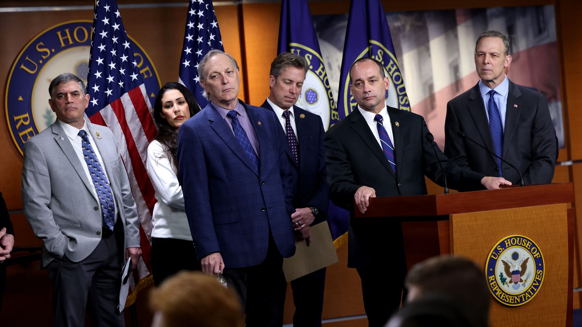 Members of the House Freedom Caucus at a press conference in a wood-paneled TV studio with a podium and several flags.