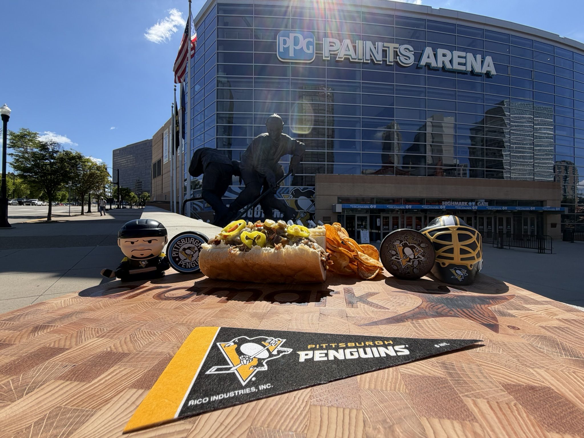Wide view of the PPG Paints Arena glass facade with an American flag flying and a hockey statue in front. In the foreground are Penguins merchandise, a large topped sandwich, and logo mats.