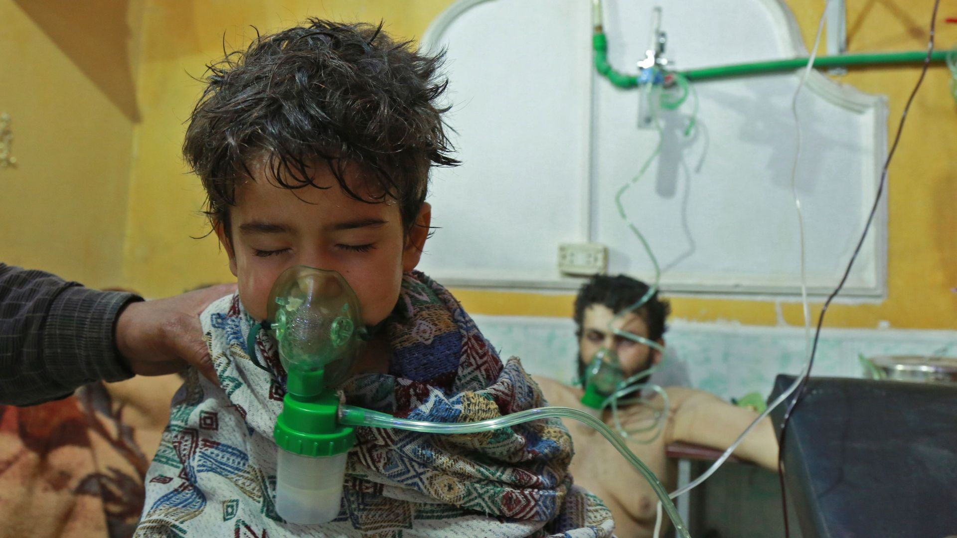 A child uses a green oxygen mask, while wrapped in a patterned blanket. A min sits behind him using a mask too. The wall is yellow.