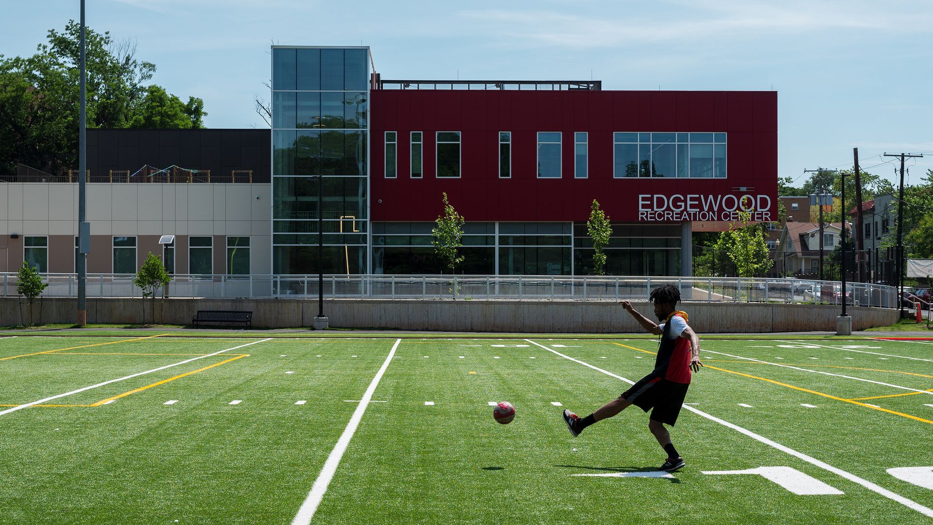 A person kicks a soccer ball on a turf field with the Edgewood Recreation Center building in the background