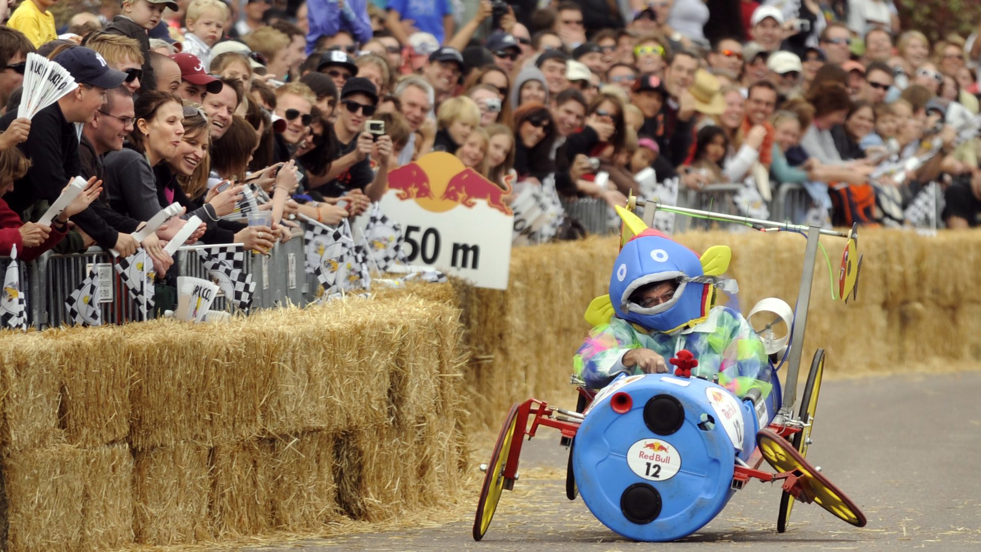 Crowded outdoor race with a racer in a blue fish costume riding a colorful, barrel-shaped soapbox car near straw bales and cheering spectators holding checkered flags.