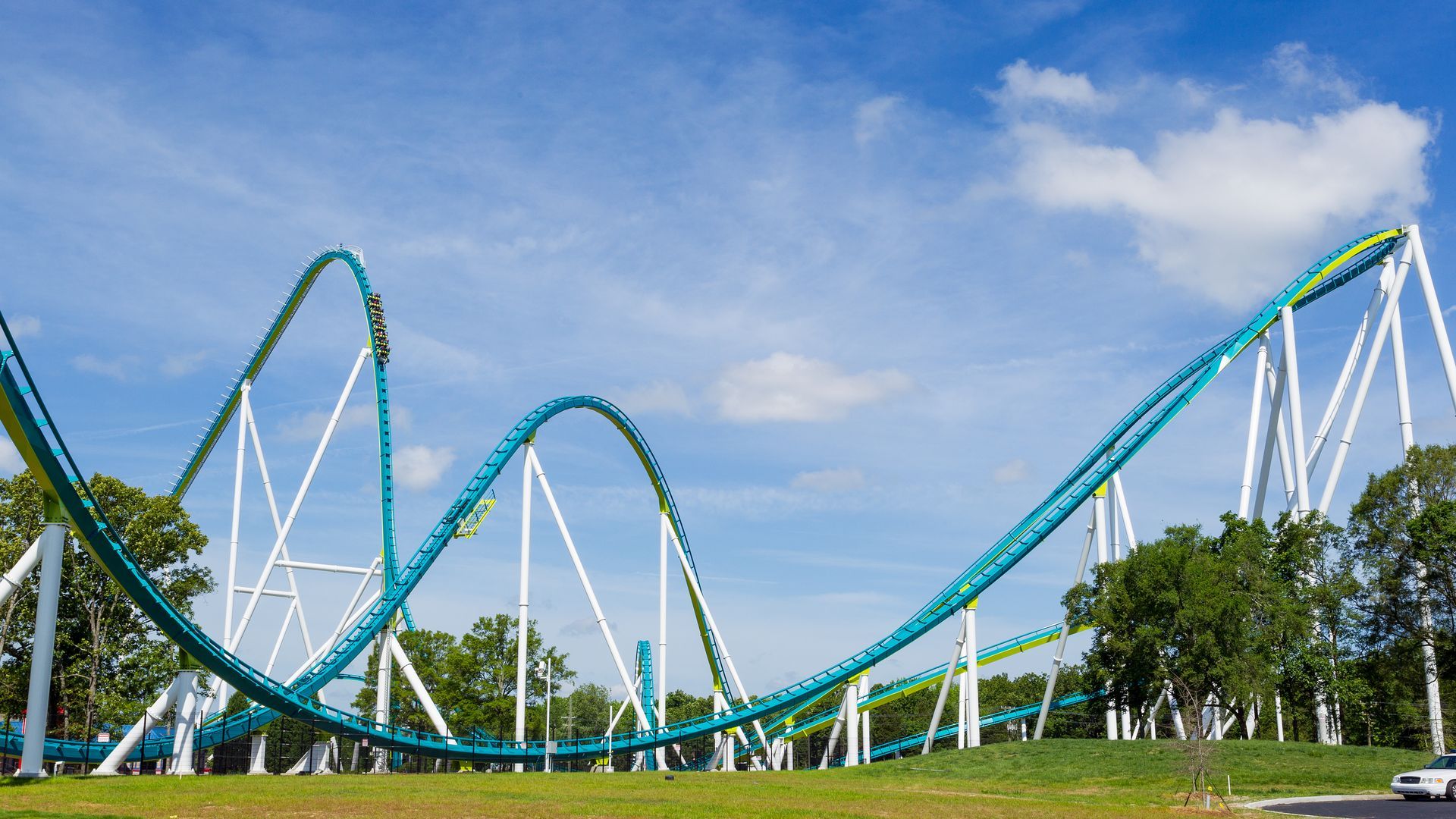 Teal roller coaster arching over white supports against a blue sky; riders in a car, with green grass and trees in the foreground.