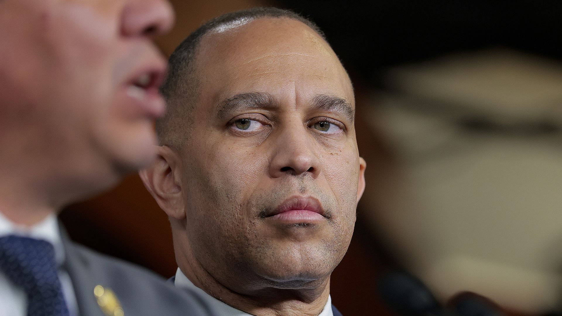 Close-up of a serious man in a navy suit and light blue tie, seated at a podium with microphones; a second man in a gray suit appears blurred in the foreground.
