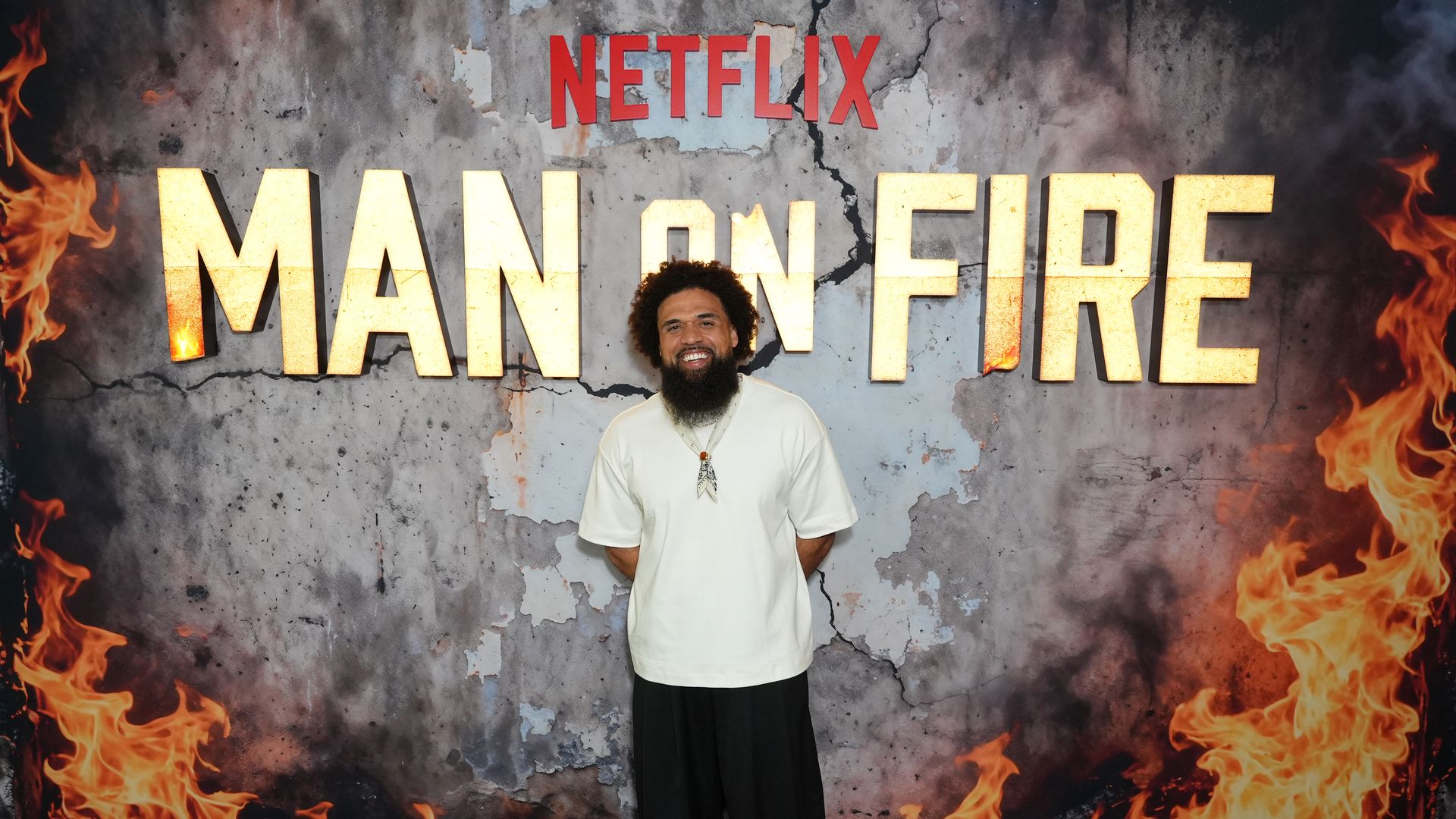 Bearded man in a white shirt and necklace smiles at the camera, standing before a cracked wall mural with flames and giant letters reading "MAN ON FIRE" and the red "NETFLIX" logo above.