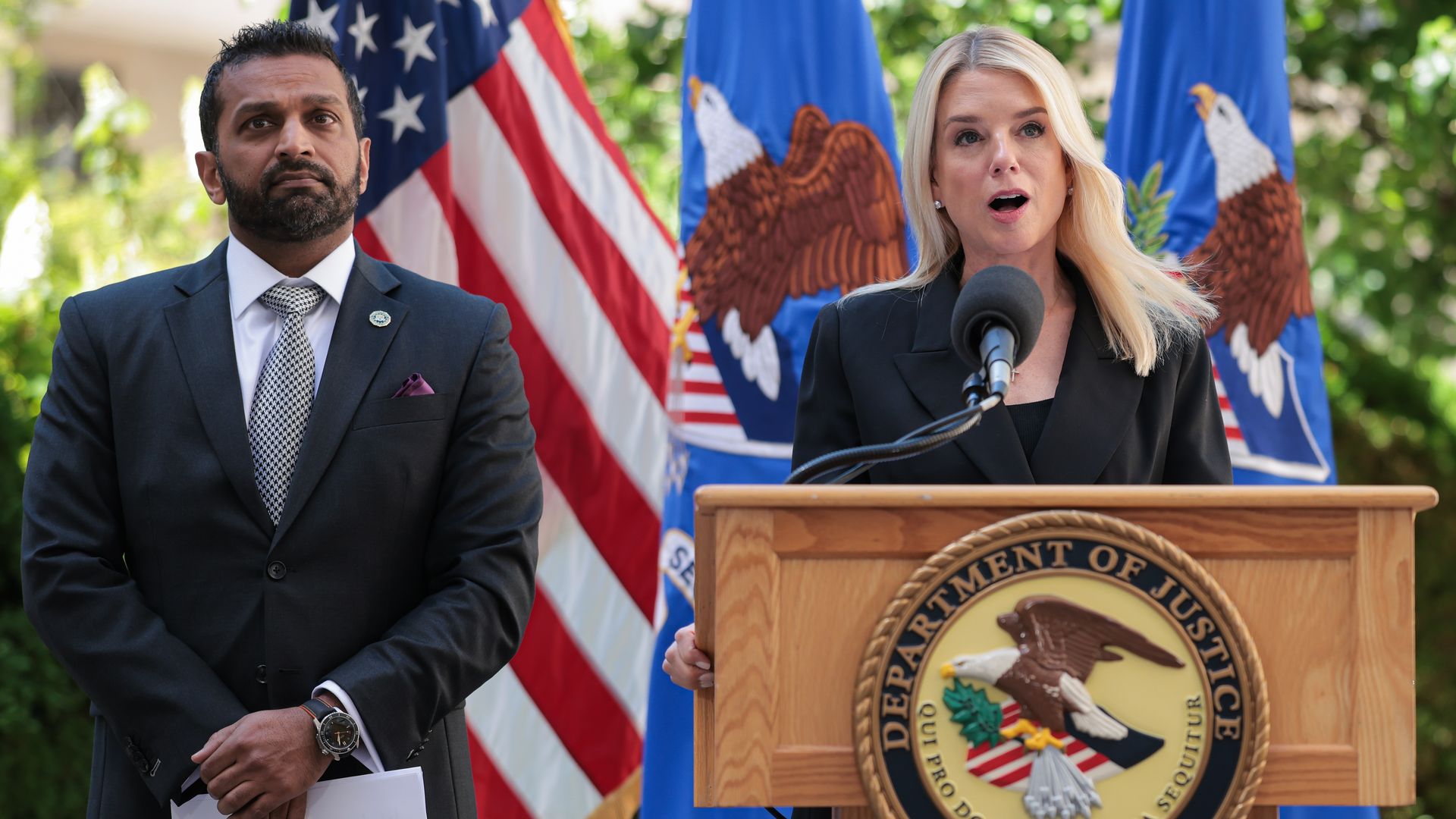 A woman with blonde hair in a black blazer speaks at a Department of Justice podium, a man in a dark suit stands beside her holding papers, with American flags and DOJ banners behind them.