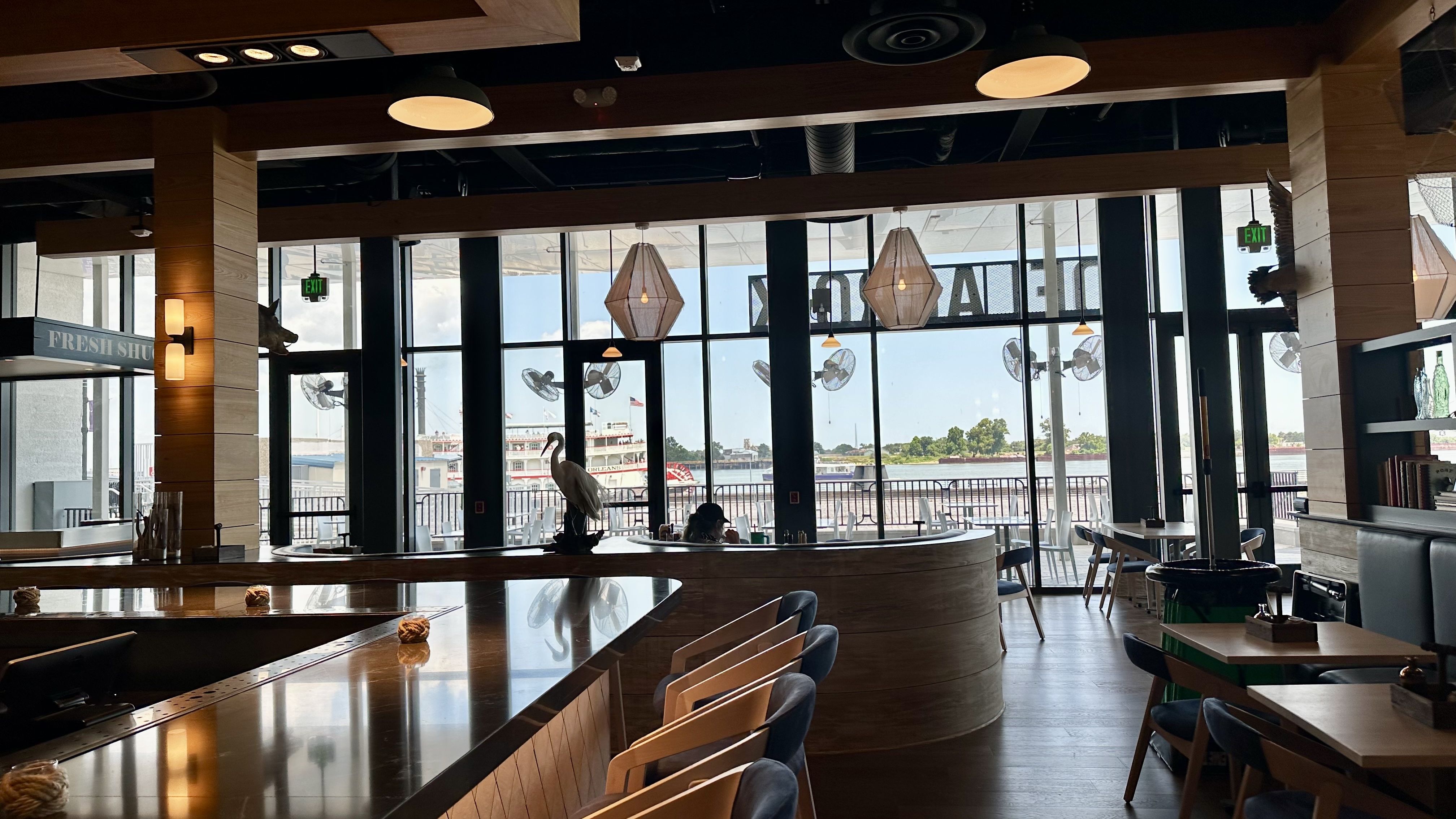 Interior of a restaurant with wooden furniture, blue chairs, hanging pendant lights, and large windows overlooking a river and a paddlewheel boat outside under a blue sky.