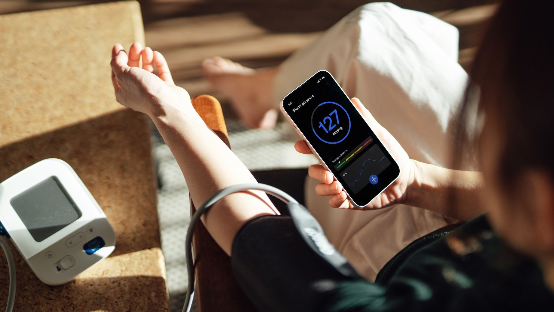 Person with a blood pressure cuff on their forearm holds a smartphone showing a circular blue reading 127 mmHg; a home BP monitor sits on a counter nearby in warm sunlight.