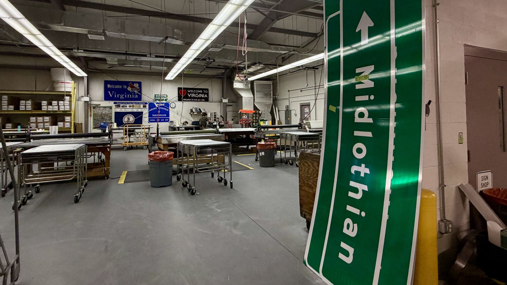 Industrial workshop with exposed ducts and bright fluorescent lights. A tall green sign with white arrows leans on the right. In the background are workbenches, carts, and Virginia welcome signs.