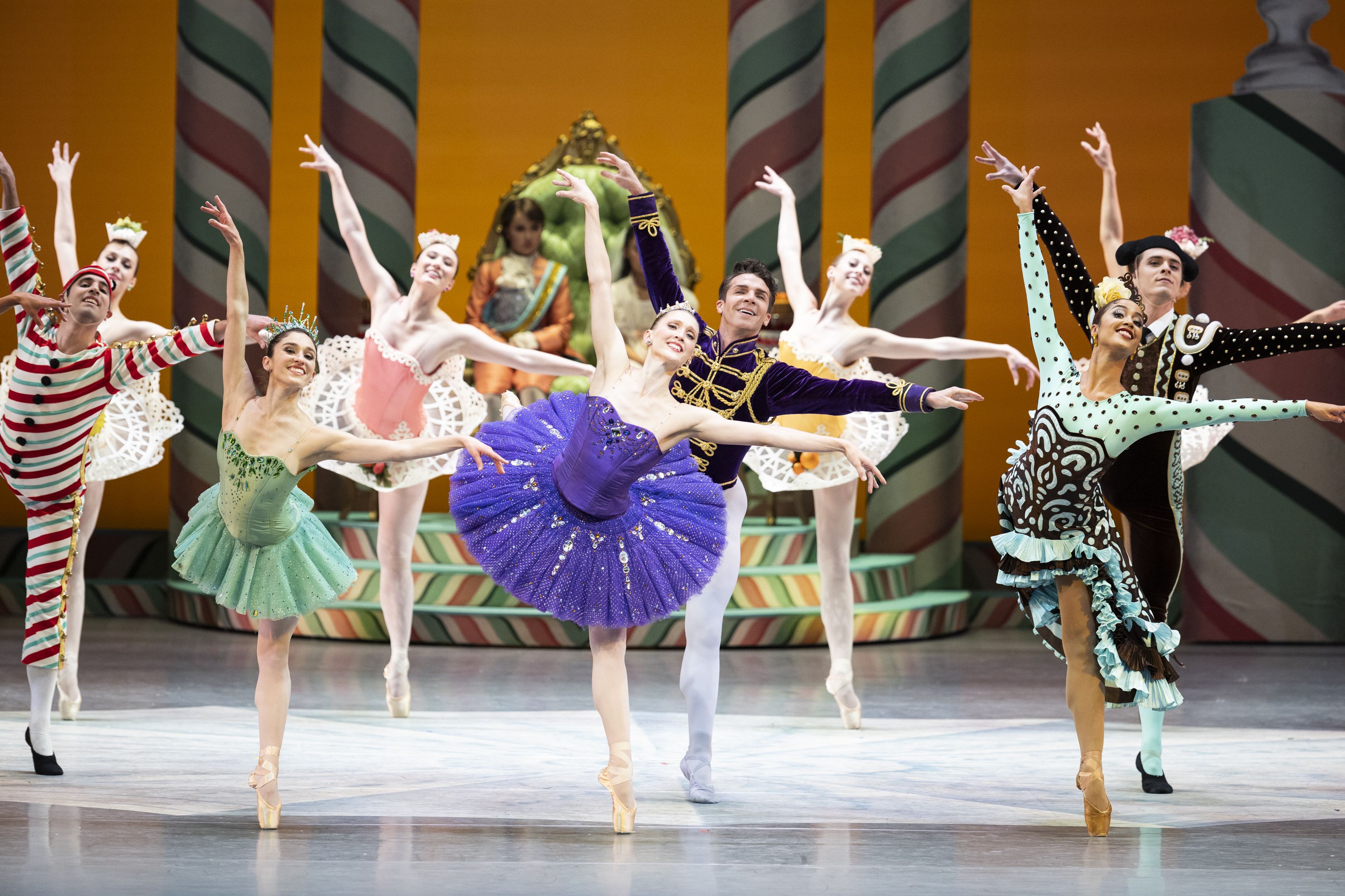 Rows of dancers lift their legs behind them in arabesques amid a candy-themed set, wearing colorful costumes.