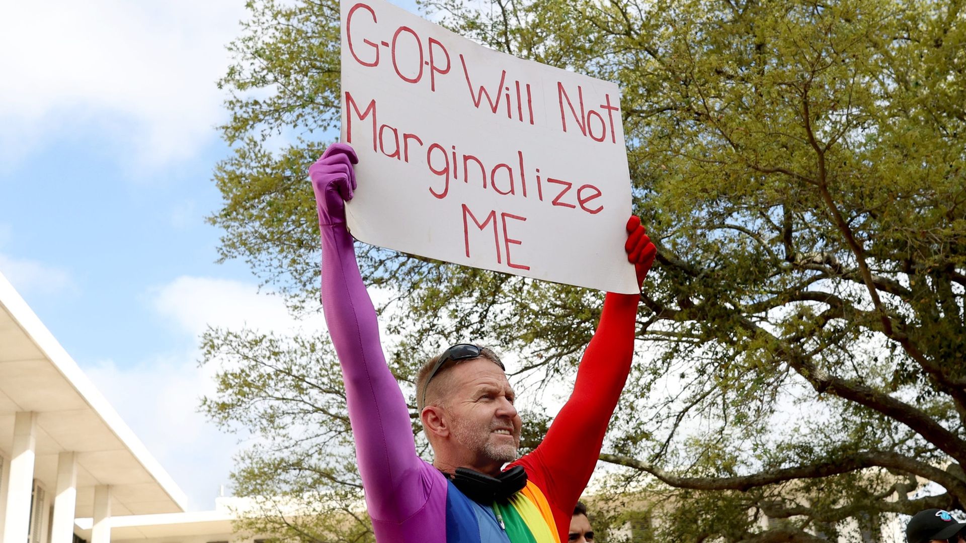 A person wearing a rainbow body suit holds up a sign saying "GOP Will not Marginalize ME"