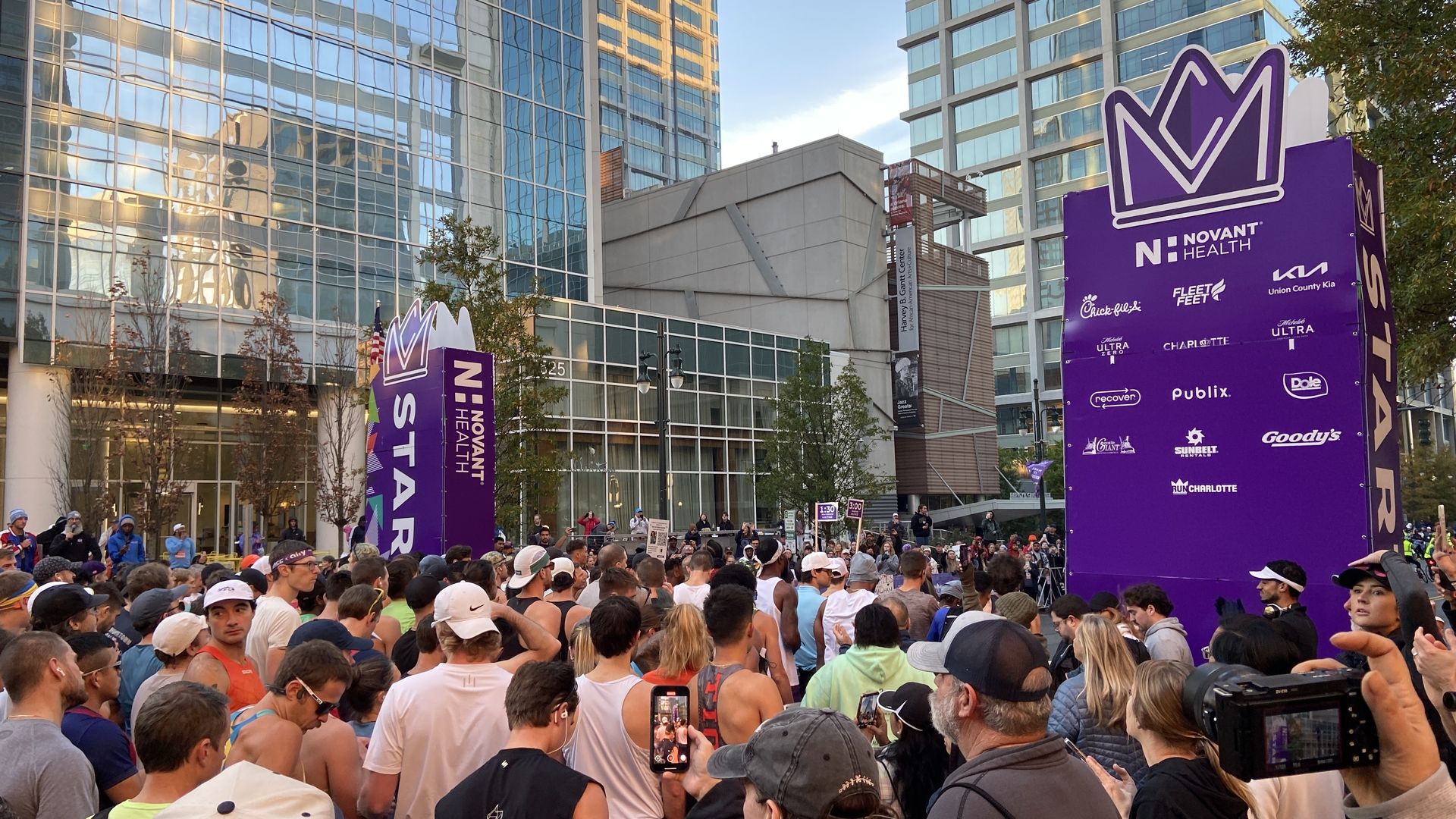 Crowd of runners at the starting line of a race in a city with tall glass buildings, next to a large purple sign with sponsors including Novant Health and Chick-fil-A.