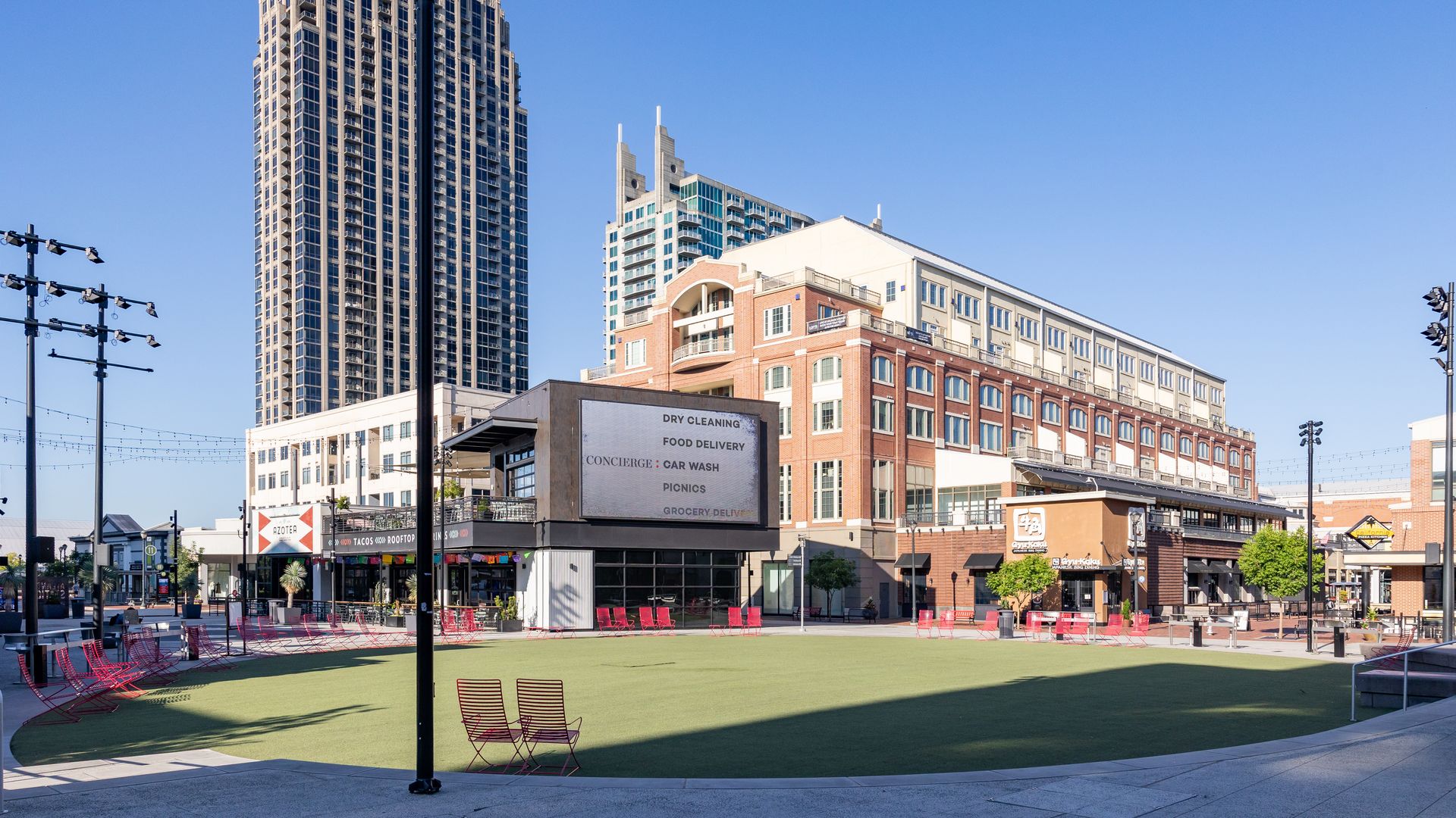 Urban plaza with red metal chairs on green artificial turf, alongside a tall gray high-rise on the left and brick/glass buildings on the right under a bright blue sky with a small moon.