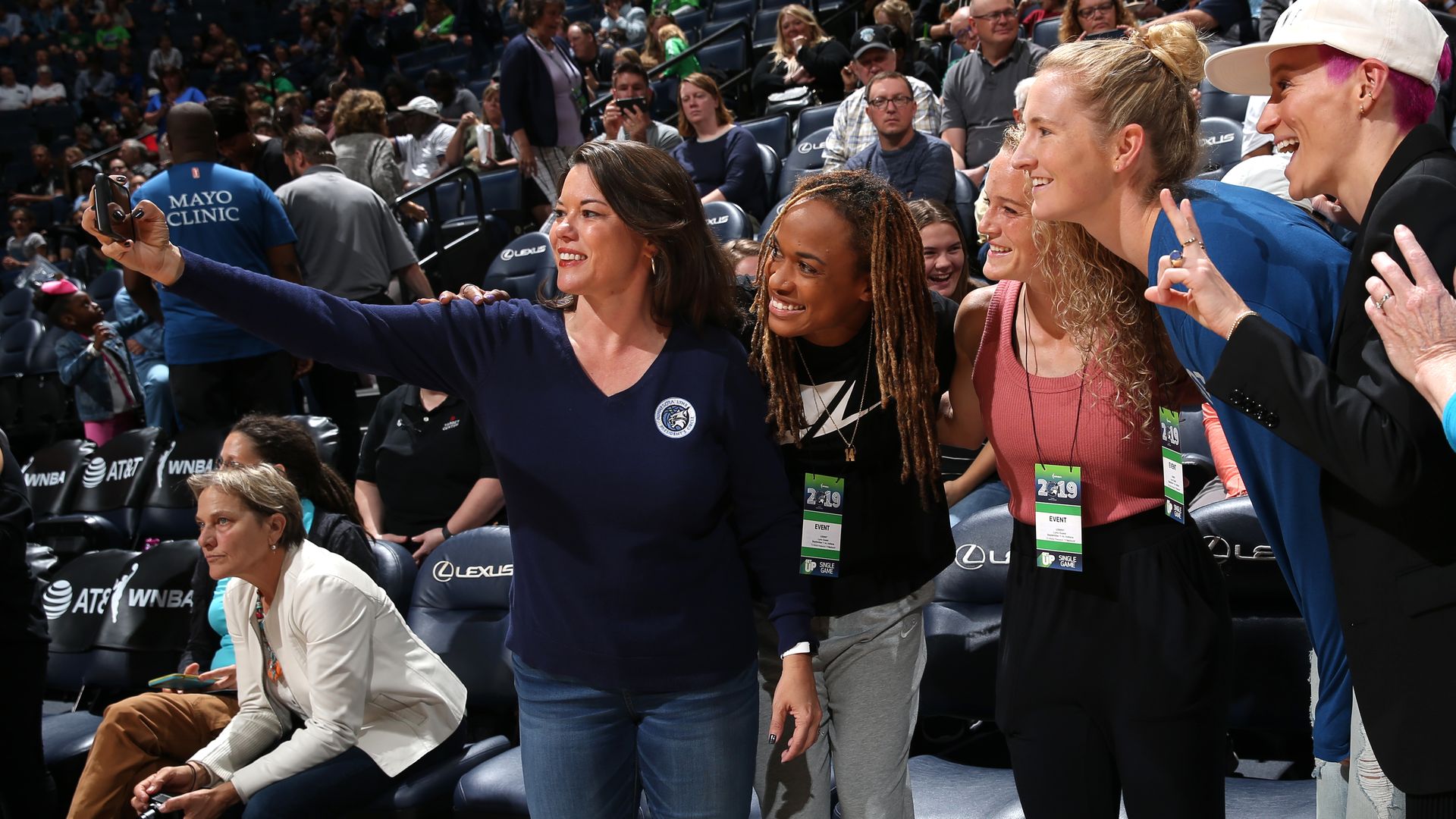 Group of smiling people pose for a selfie at a sports arena; woman in navy shirt extends arm with phone while others in pink top, blue jacket and white cap lean in; crowd in stands behind.