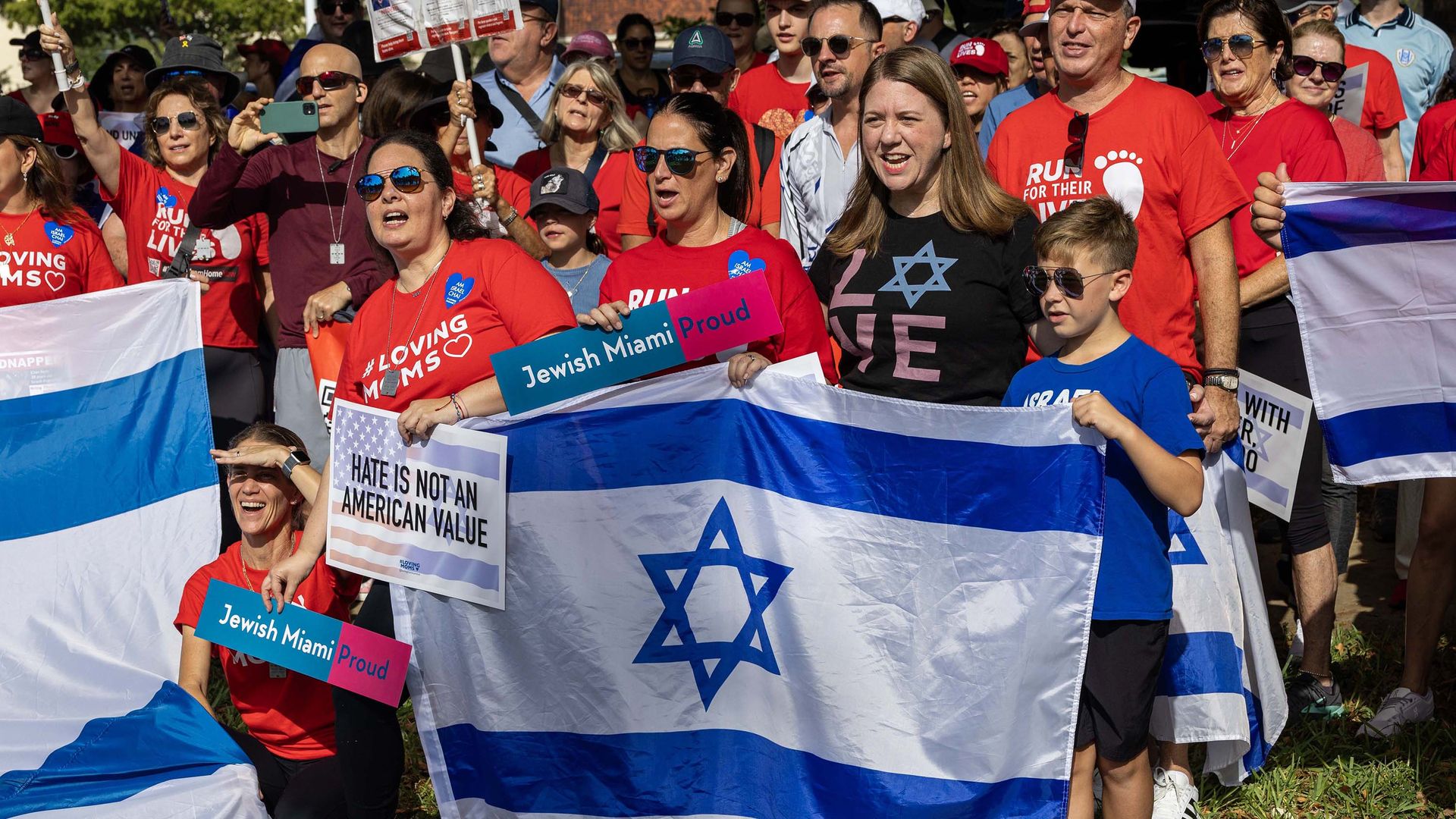 Group of people, many wearing red shirts, hold Israeli flags and signs reading "Jewish Miami Proud" and "Hate is not an American value" at an outdoor event on a sunny day.