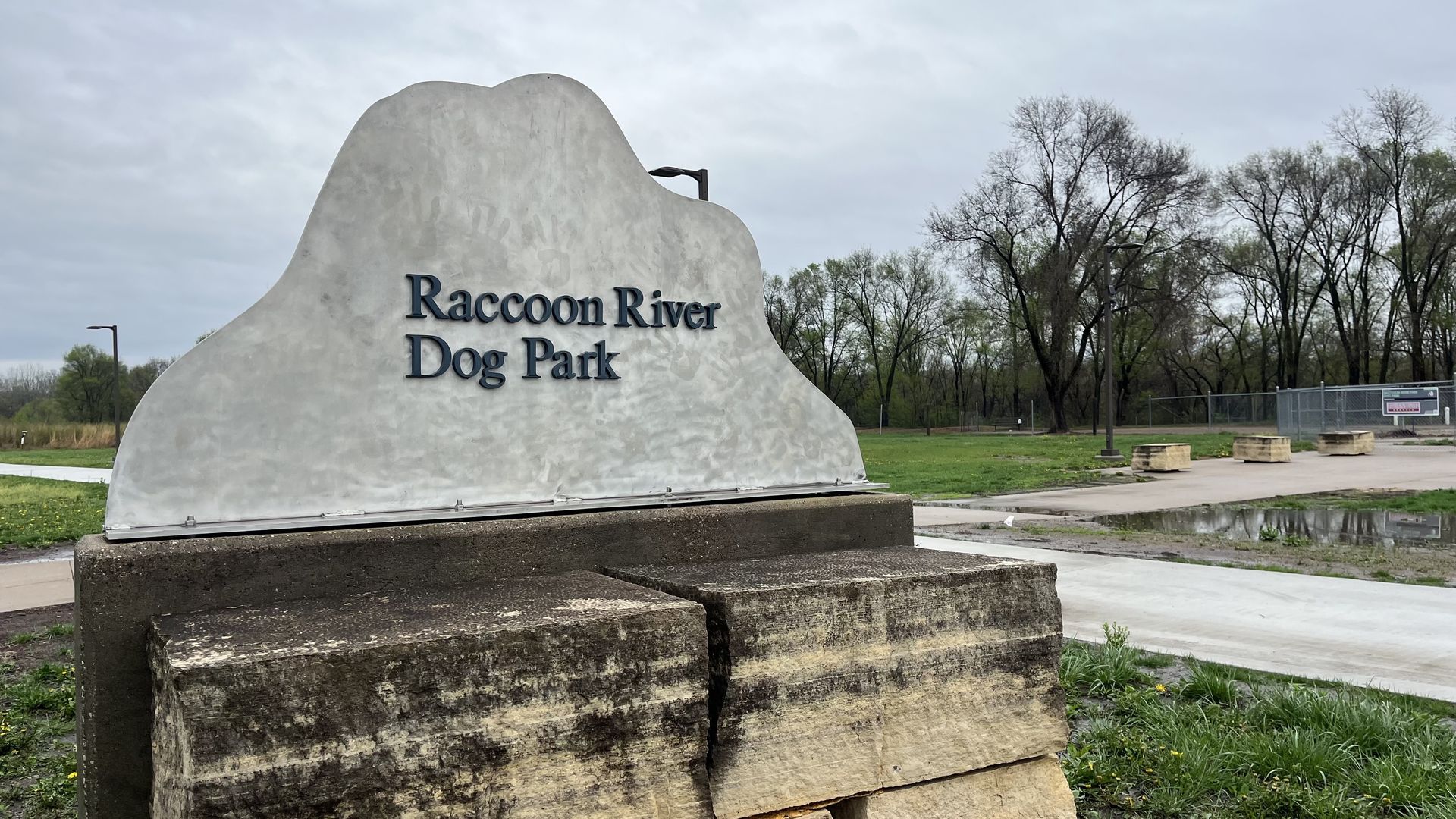 A large grey rock-like sign with dark blue lettering reads "Raccoon River Dog Park", set on a concrete pedestal near trees and a fence under a cloudy sky.