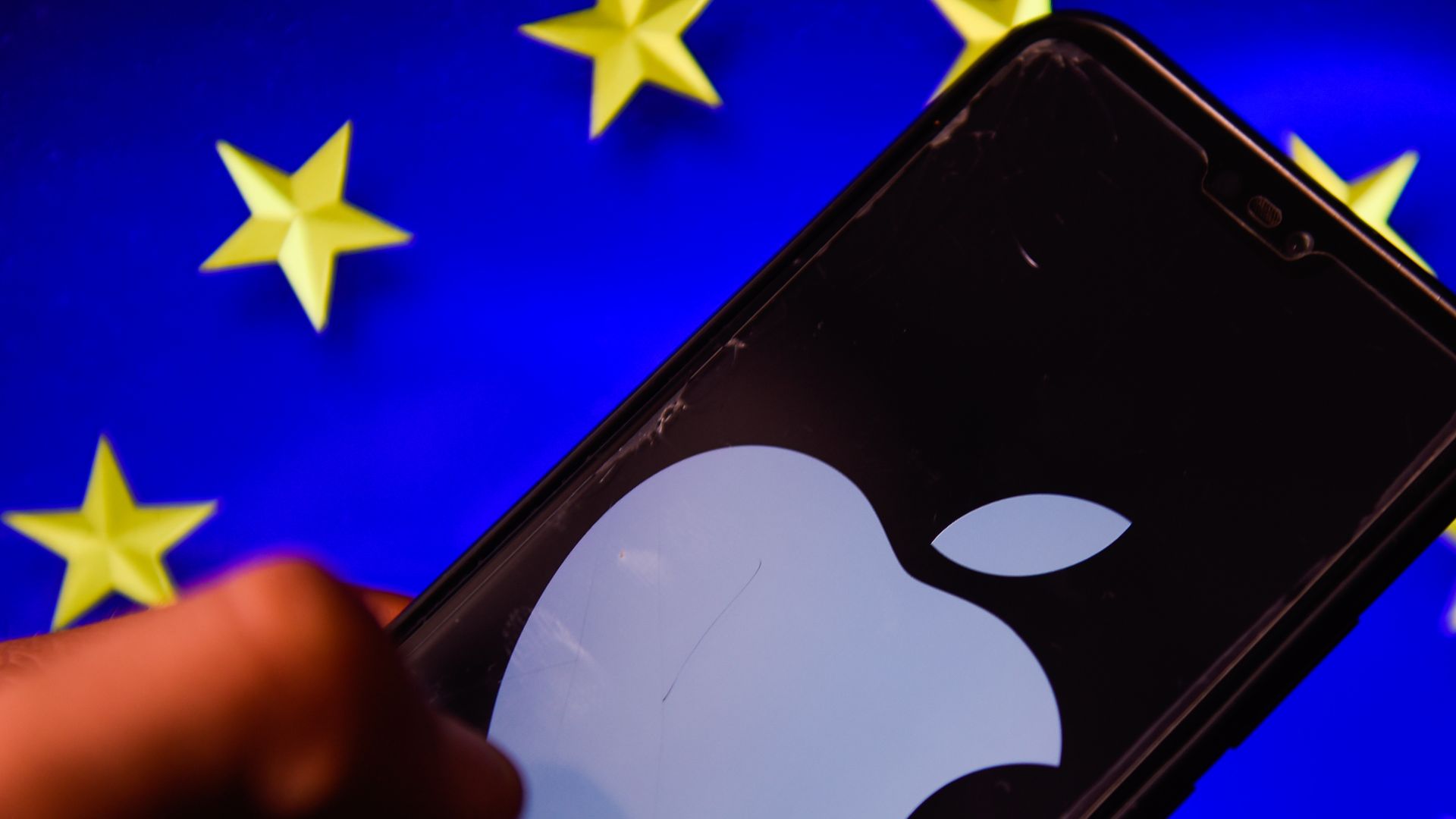 A photo illustration of a smartphone with the Apple logo displayed prominently on the screen, with a European Union flag in the background.