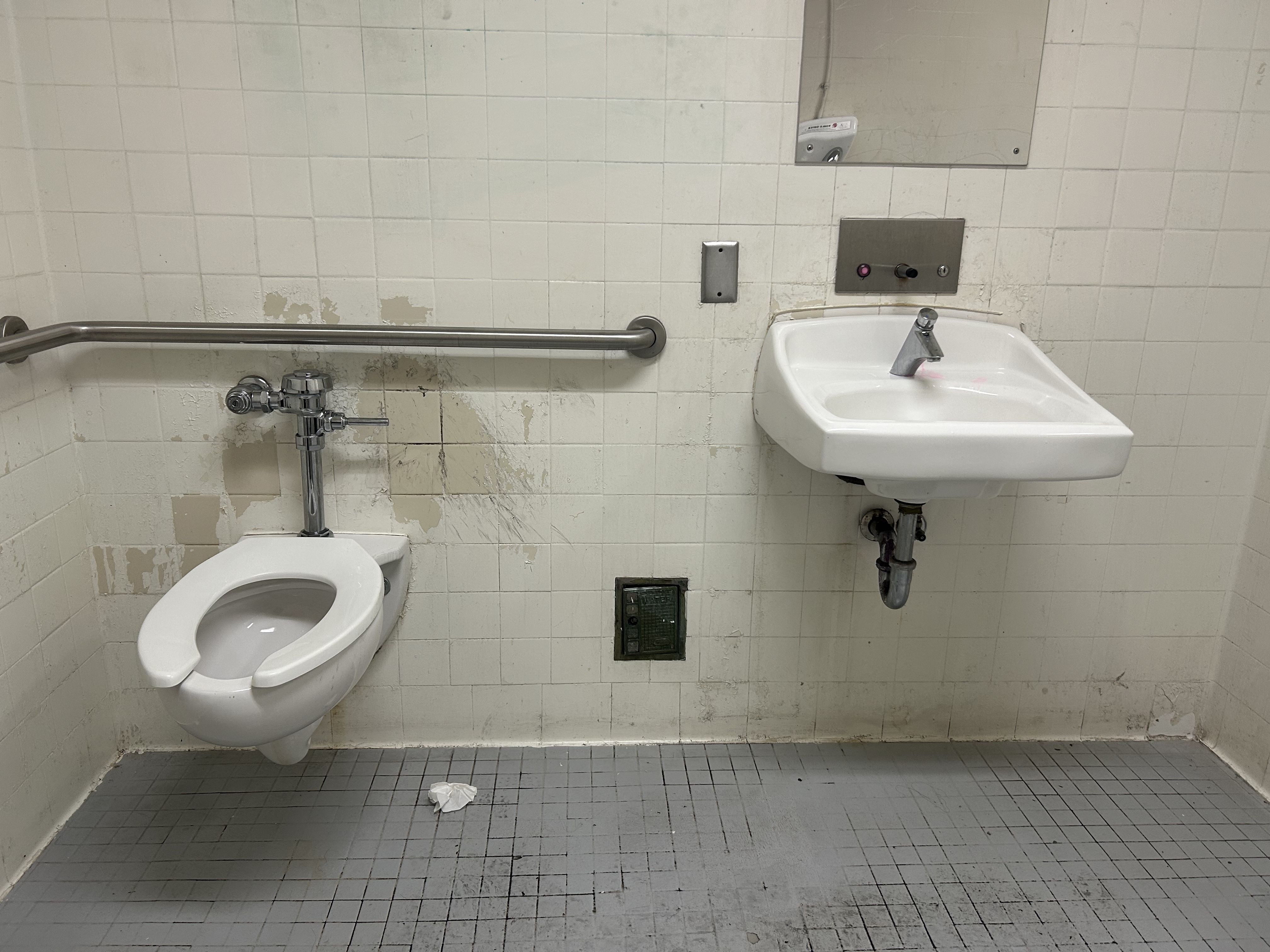 Public restroom with white tiled walls. A wall-mounted sink on the right has exposed pipes below; a left toilet appears to have no seat. A long grab bar runs along the wall, with a floor vent and dirt patches.