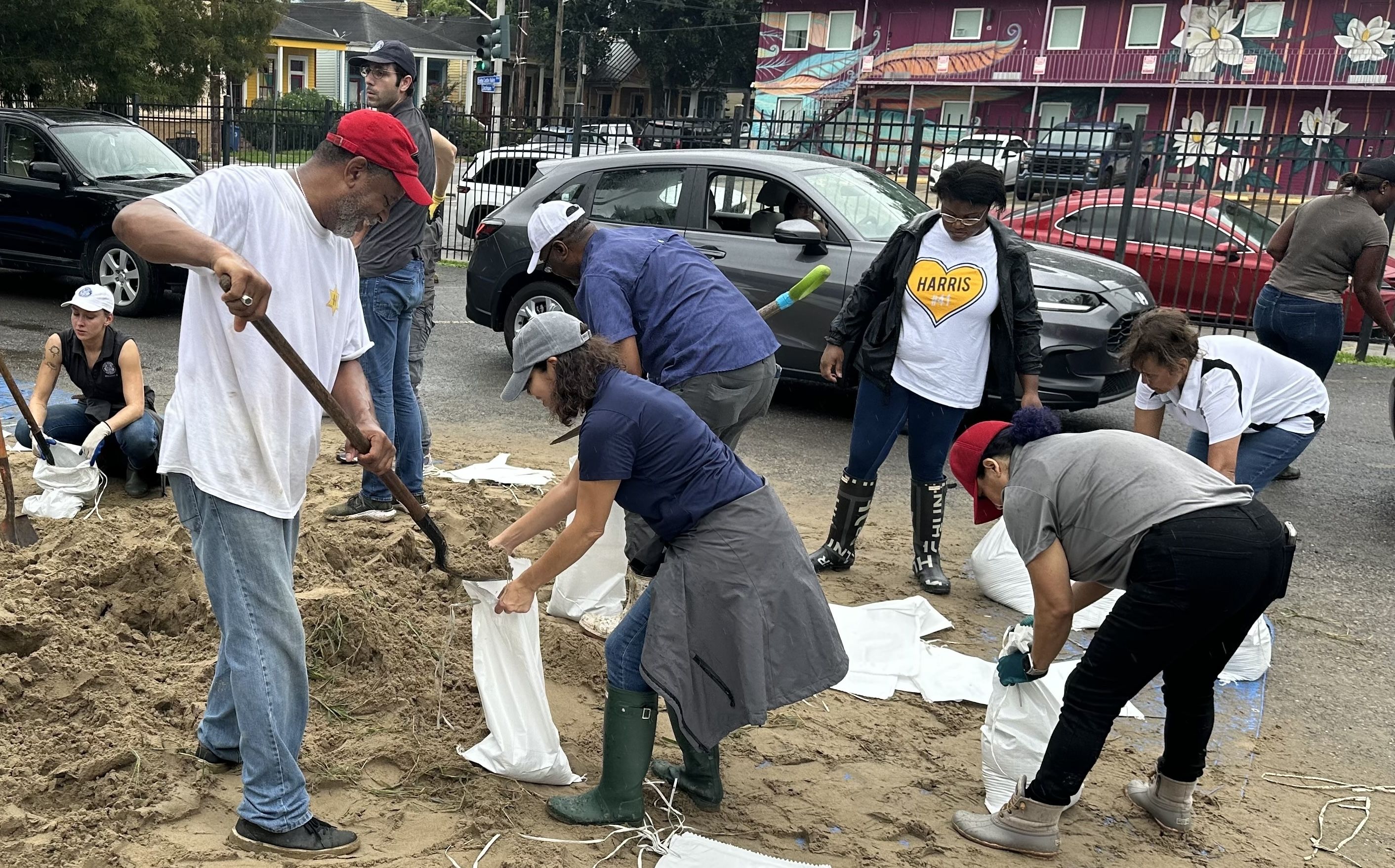 A diverse group of people outdoors filling white sandbags from a large pile, near parked cars and colorful buildings with flower murals, wearing casual clothes and boots.