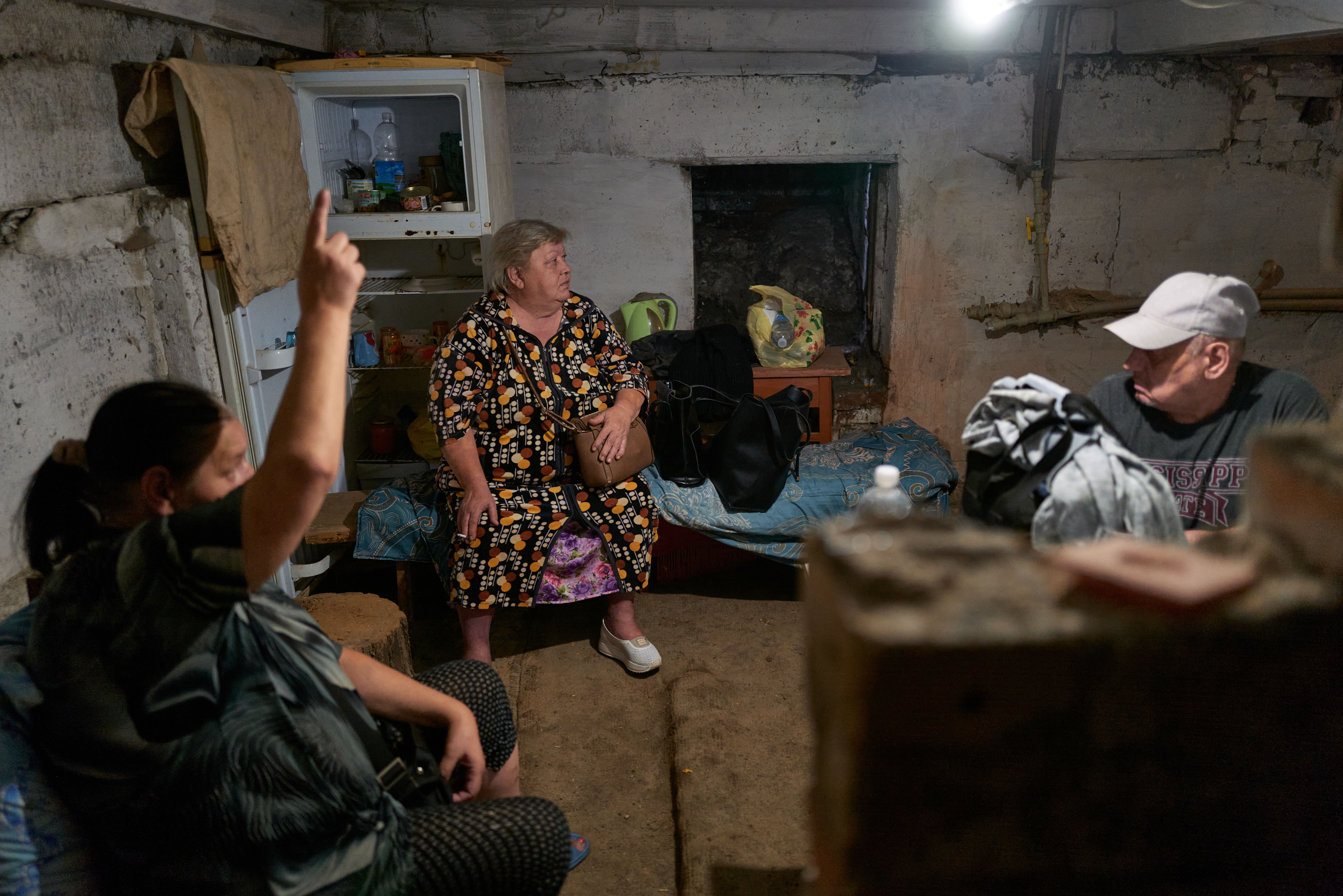 Three people in a dim, rustic basement room with concrete walls and a fridge; a woman in a patterned dress sits on a bed, another woman raises a finger, and a man in a cap looks on.