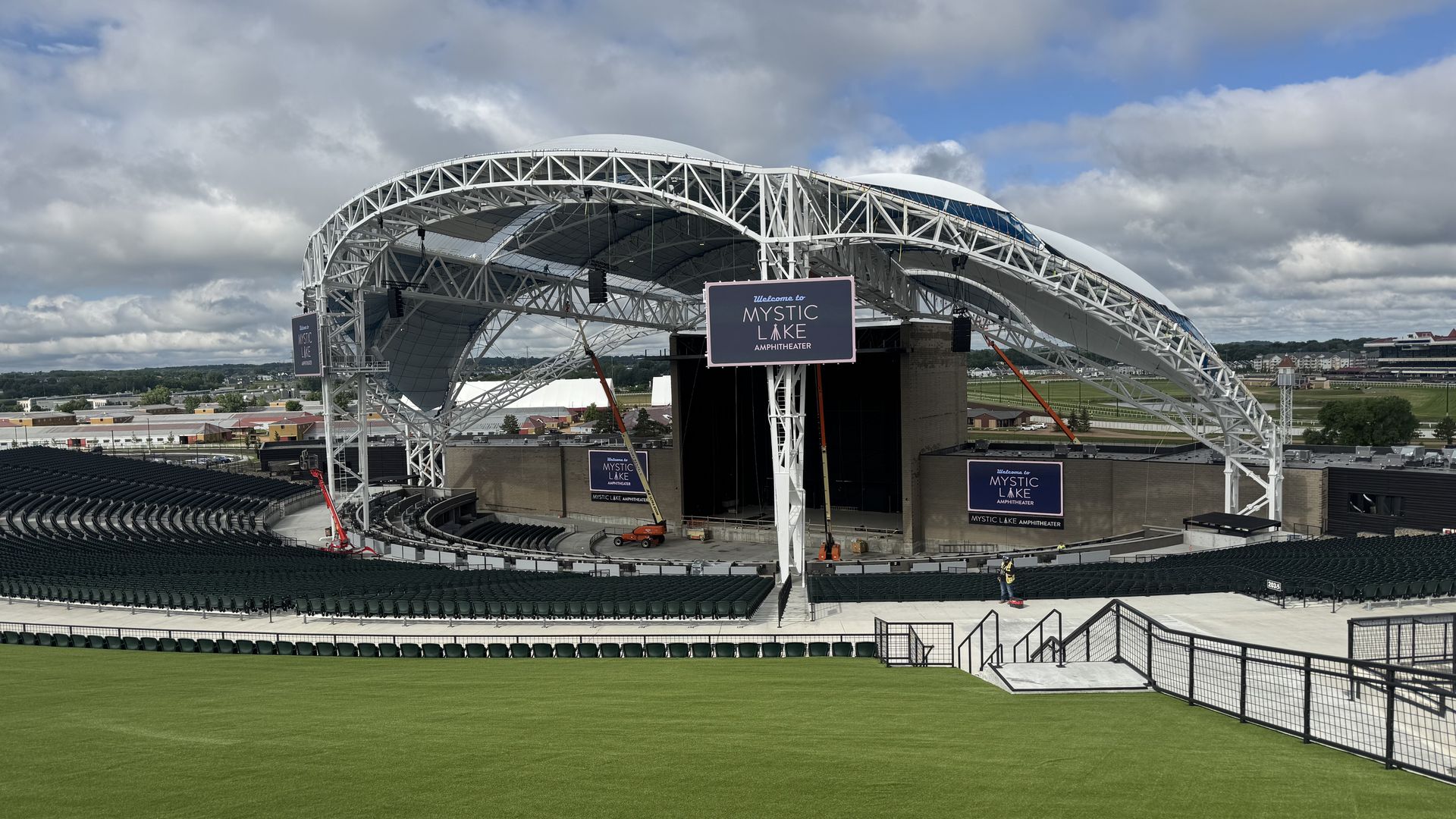 Outdoor amphitheater with a white steel arch roof, open stage and surrounding seats, green field in foreground, banners reading "Mystic Lake Amphitheater" under a cloudy blue sky.