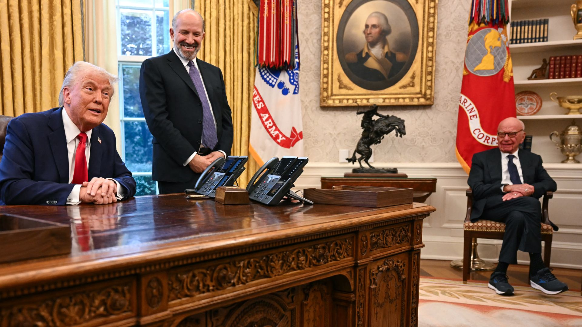 In the White House President Trump, wearing a red tie sits at an ornate wooden desk, Commerce Secretary Howard Lutnick, wearing a dark suit and Rupert Murdoch, wearing a black suite and sneakers sits on a chair near shelves.