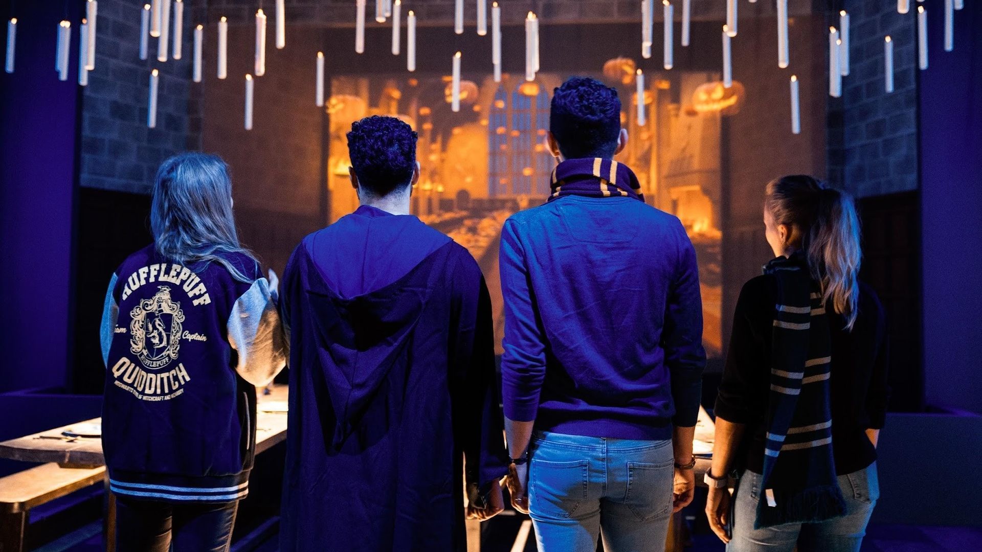 Group of five people seen from behind, wearing blue jackets (one with a Hufflepuff crest). They stand beneath hanging white cylindrical lights in a dark purple exhibition space, facing a warm orange glow.