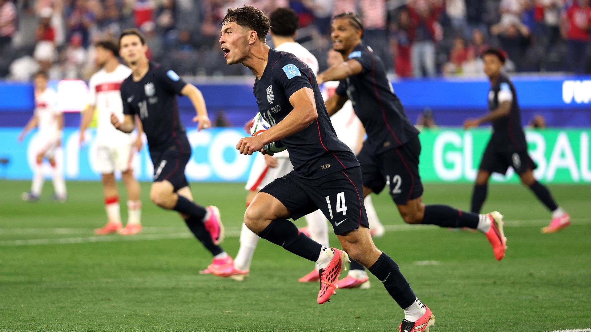 Action shot of male soccer players in dark uniforms sprinting on a green pitch; central player wearing number 14 holds a multicolored ball as teammates race alongside, crowded stadium in background.