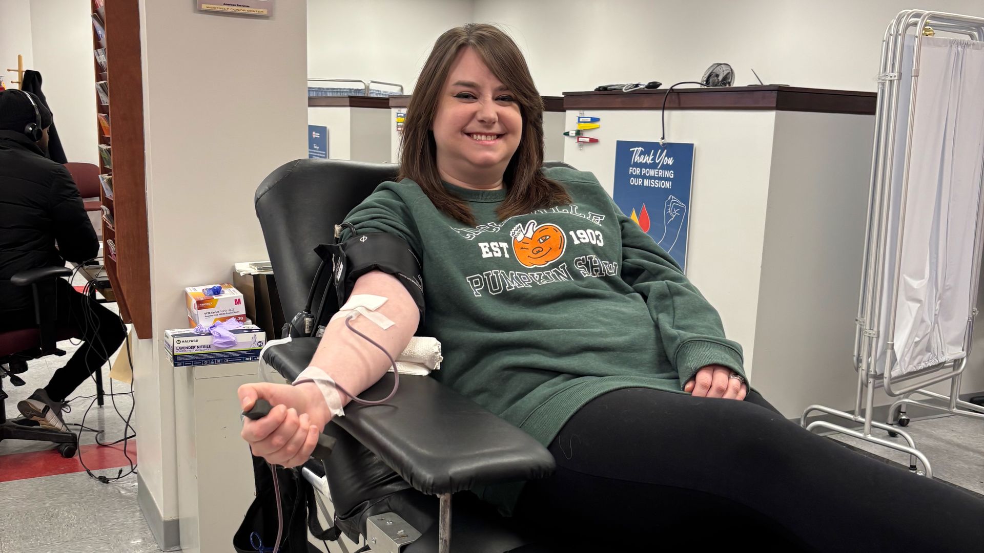 Alissa smiles while donating blood, wearing a green sweatshirt with an orange pumpkin logo, lying on a black donation chair in a clinical room.