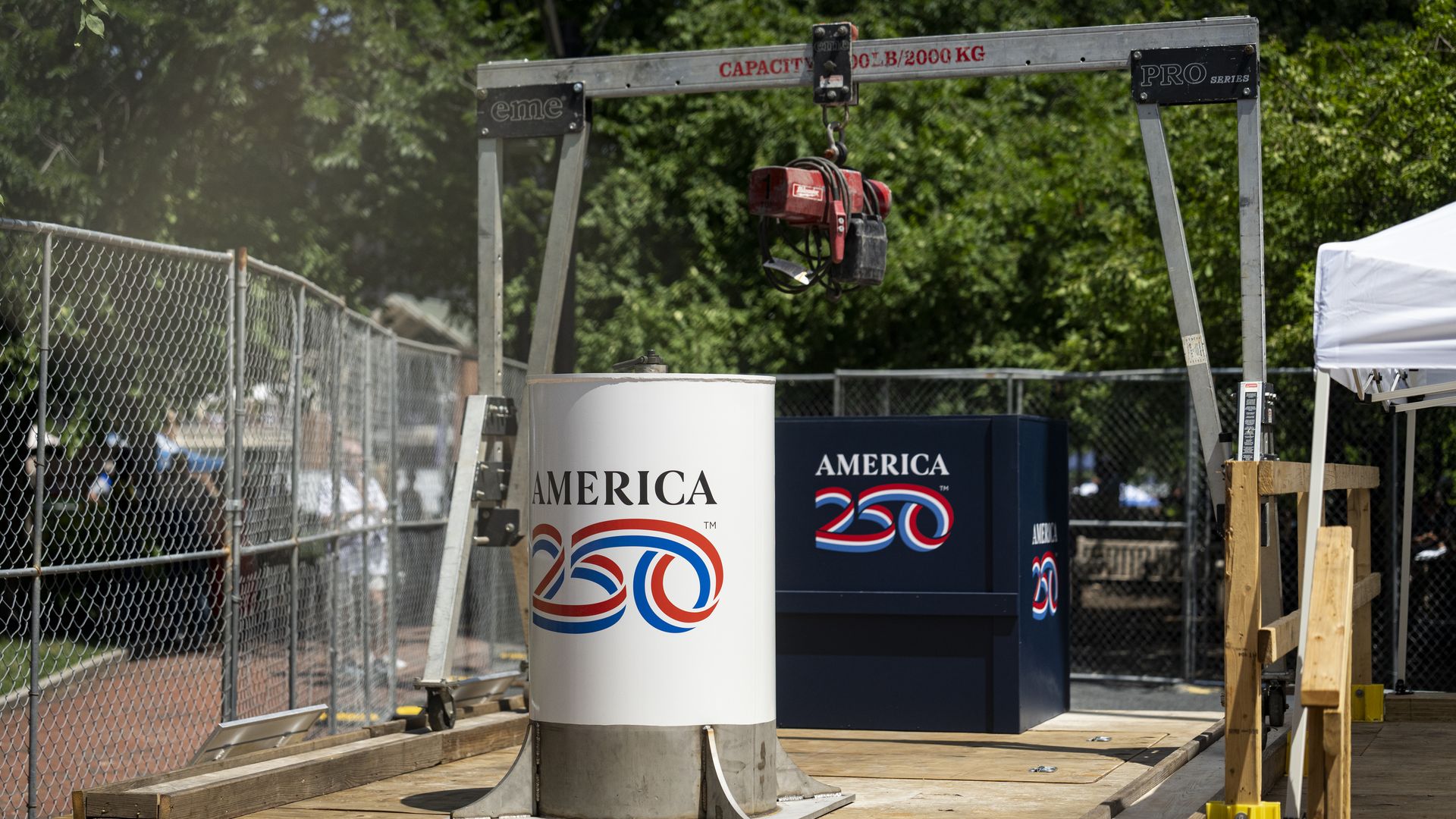 Industrial outdoor scene: grey metal gantry crane with a red hoist, chain-link fence left, white and blue American-themed barrels, wooden platform, and a white tent amid green trees.