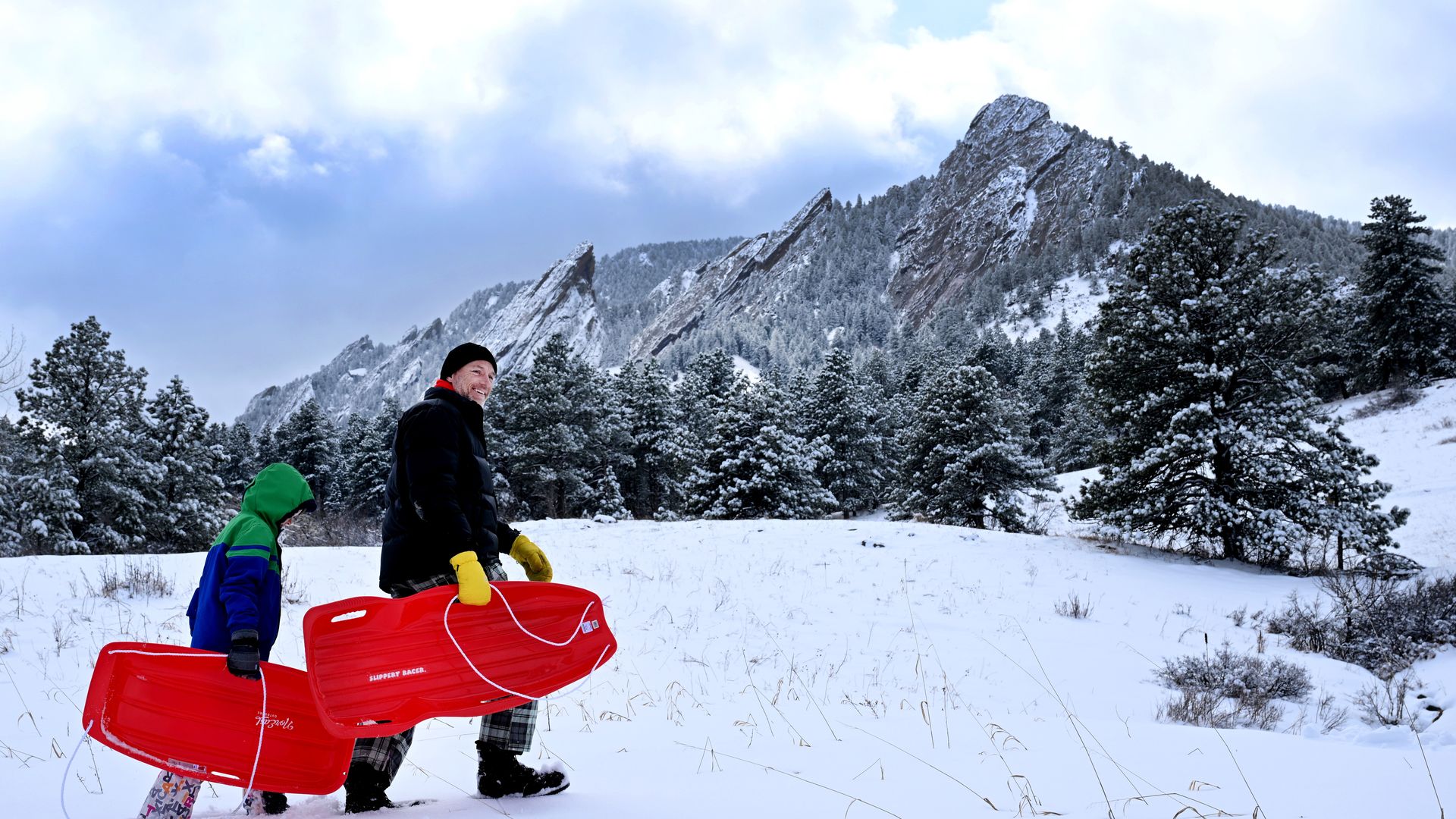 Two people in winter clothes holding red sleds walk through a snowy field with snow-covered pine trees and jagged mountains in the background under a cloudy sky.