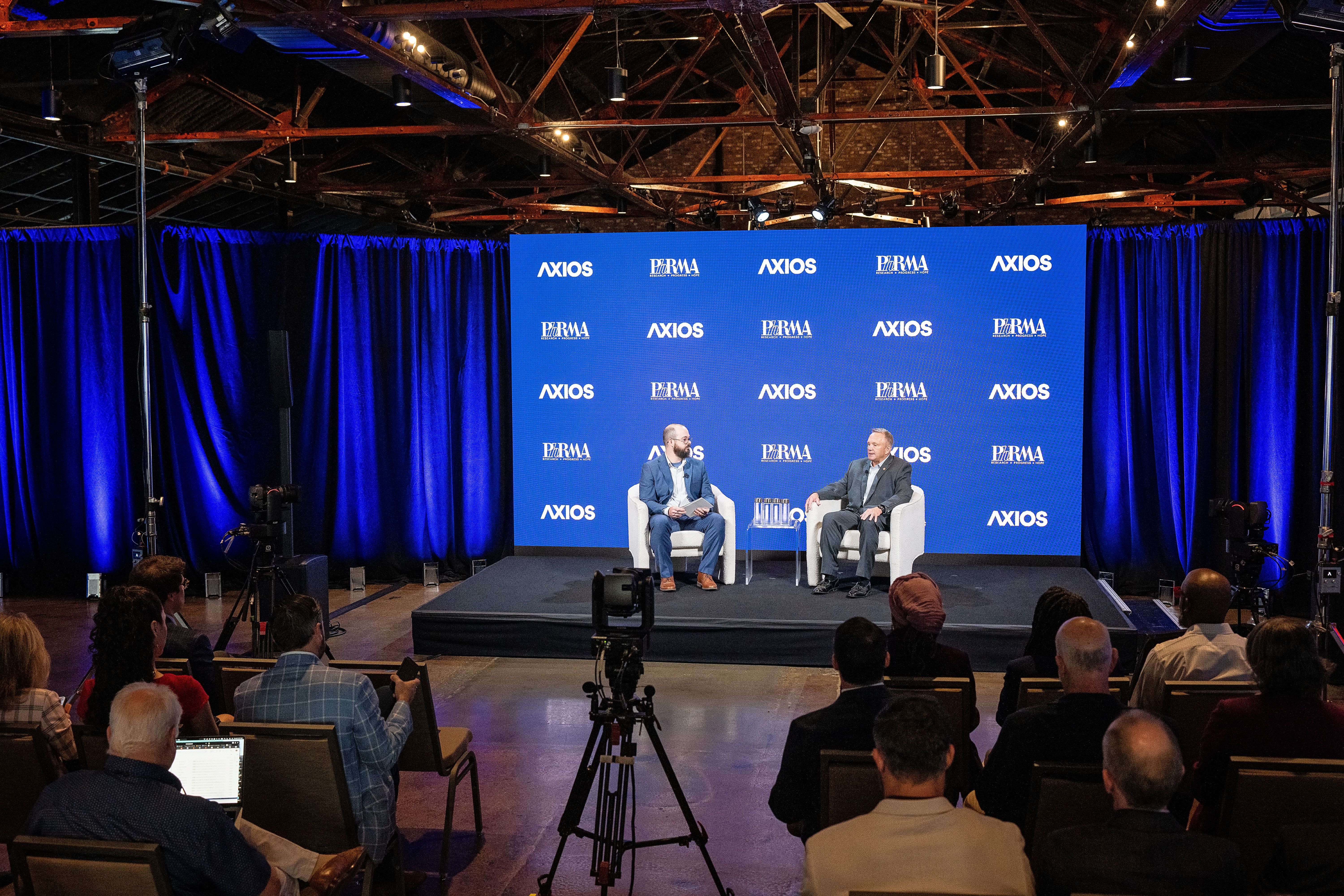 Two men sit in chairs on a small stage against a blue backdrop filled with Axios logos, flanked by blue curtains, as cameras and a seated audience look on in a dim, industrial venue.