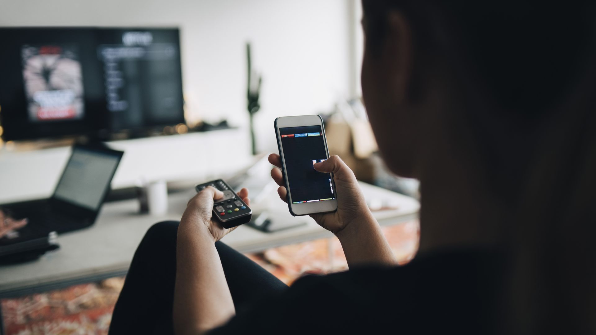 In this image, a person sits and holds a phone up to a TV mounted on the wall.