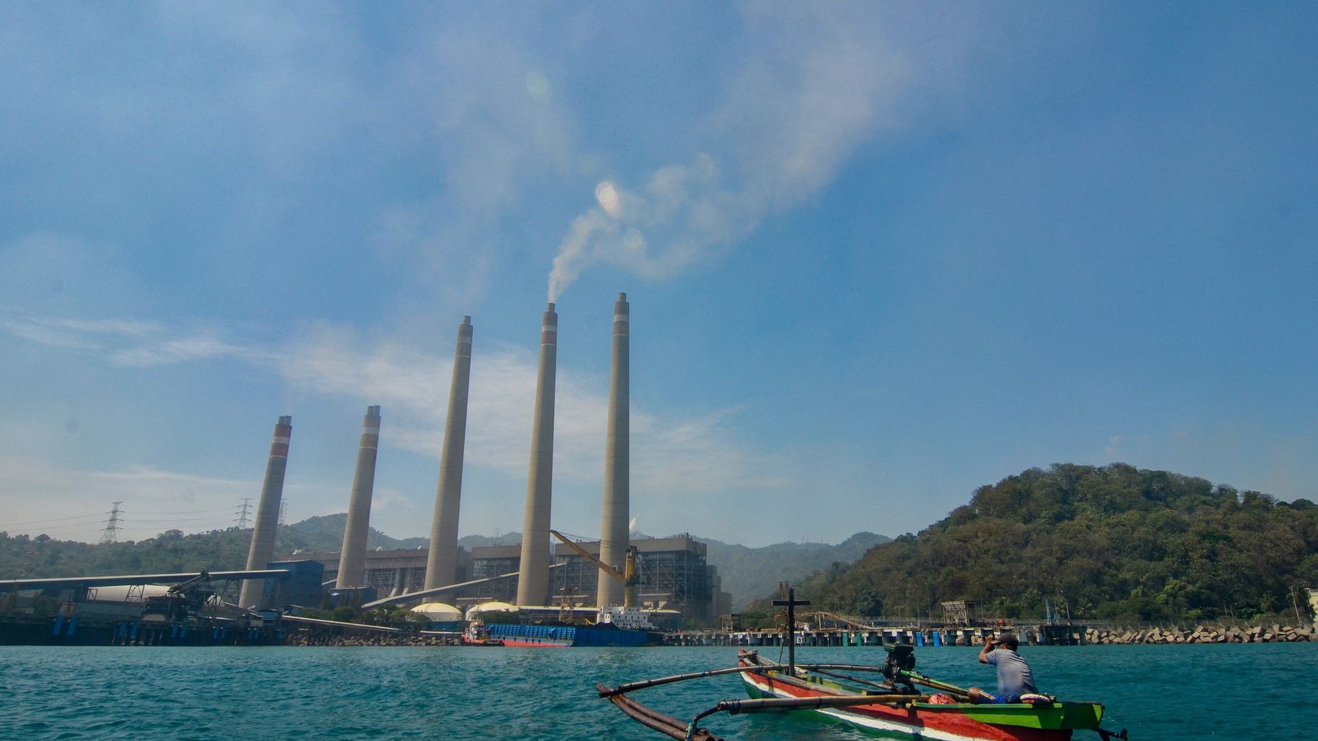This photo taken on September 22, 2021 shows fishermen on their boat as smoke rises from chimneys at the Suralaya coal power plant in Cilegon, Indonesia. Photo by RONALD SIAGIAN/AFP via Getty Images