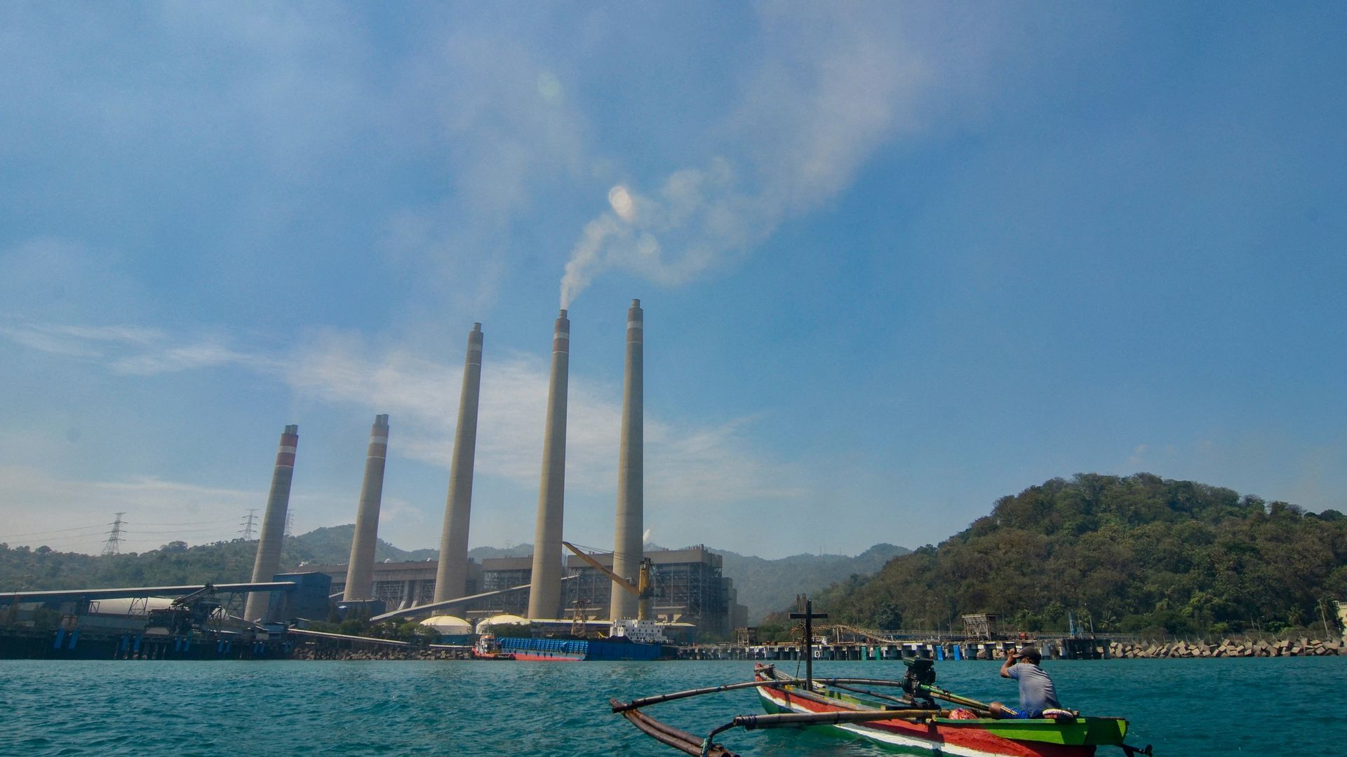 This photo taken on September 22, 2021 shows fishermen on their boat as smoke rises from chimneys at the Suralaya coal power plant in Cilegon, Indonesia. Photo by RONALD SIAGIAN/AFP via Getty Images