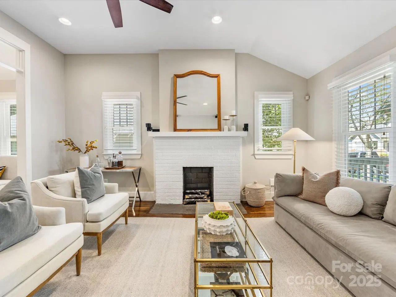 Bright living room with white brick fireplace, wooden-framed mirror, two beige armchairs with gray pillows, gray sofa, glass coffee table, and large windows with white blinds.
