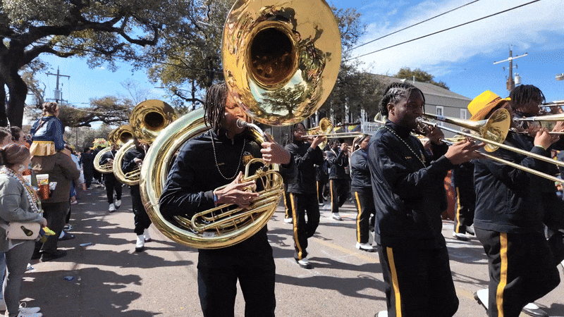 Marching band in black and gold uniforms playing brass instruments, including sousaphones and trombones, parading down a street lined with spectators on a sunny day.