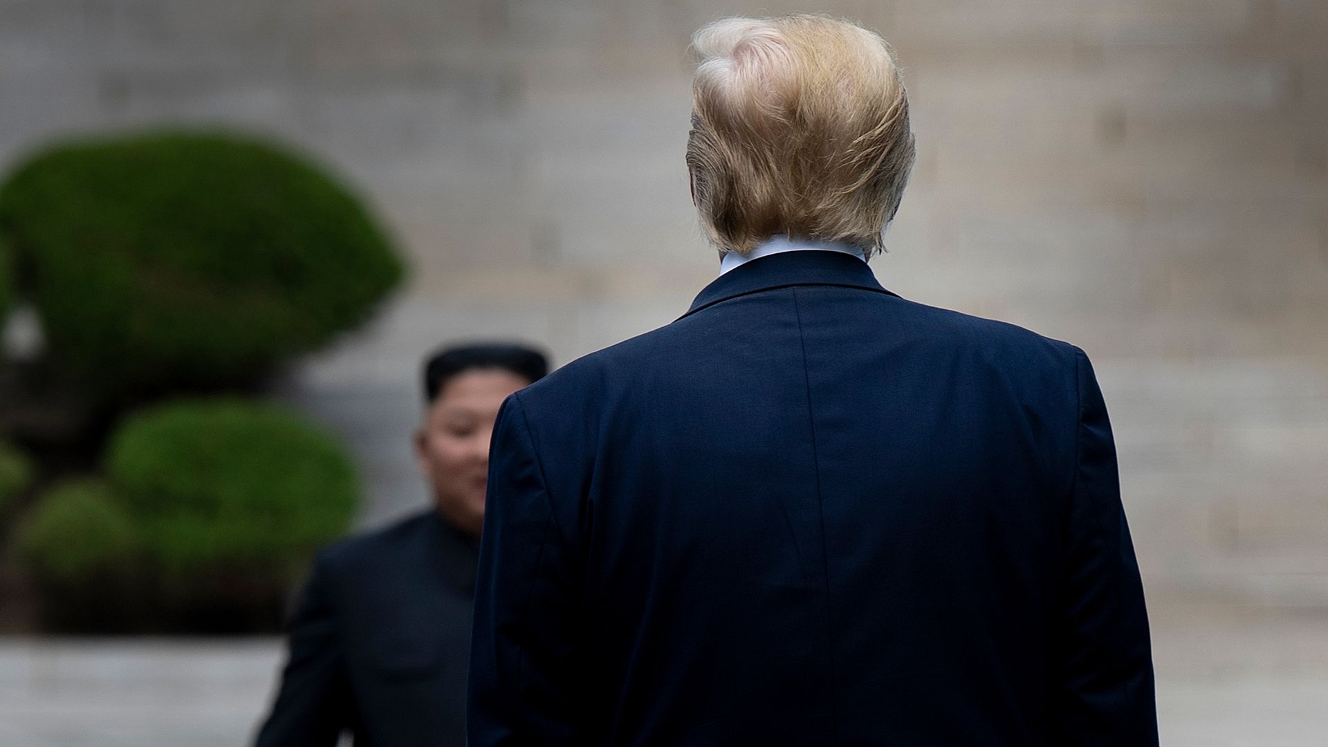 In this image, the back of Trump's head and silhouette is visible as he faces Kim Jong Un, who smiles at the camera. They are walking towards each other.