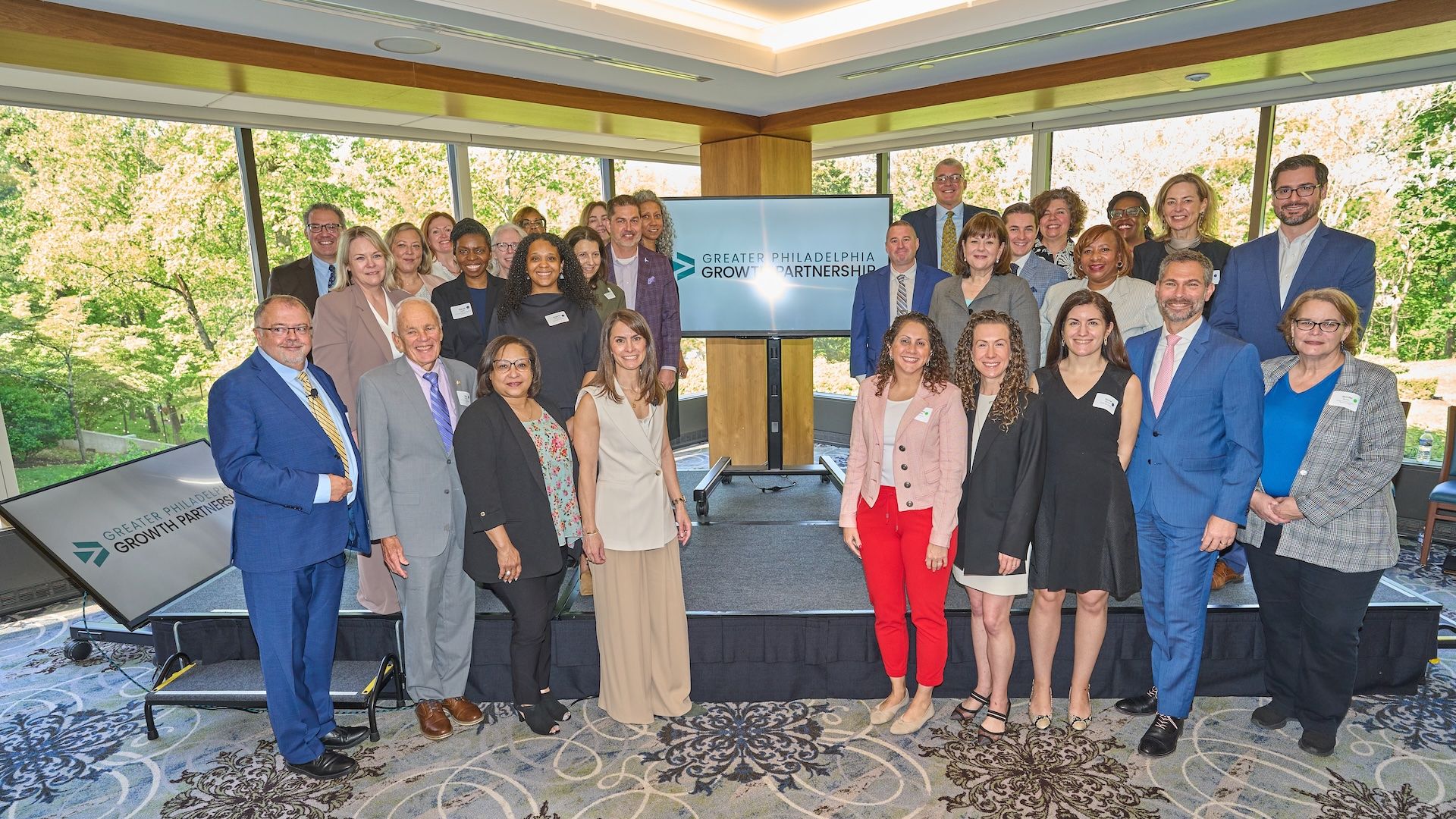 Diverse group of professionals in a sunlit conference room pose around a central screen that reads "Greater Philadelphia Growth Partnership"; they wear business attire (suits, blazers) with greenery visible through large windows.
